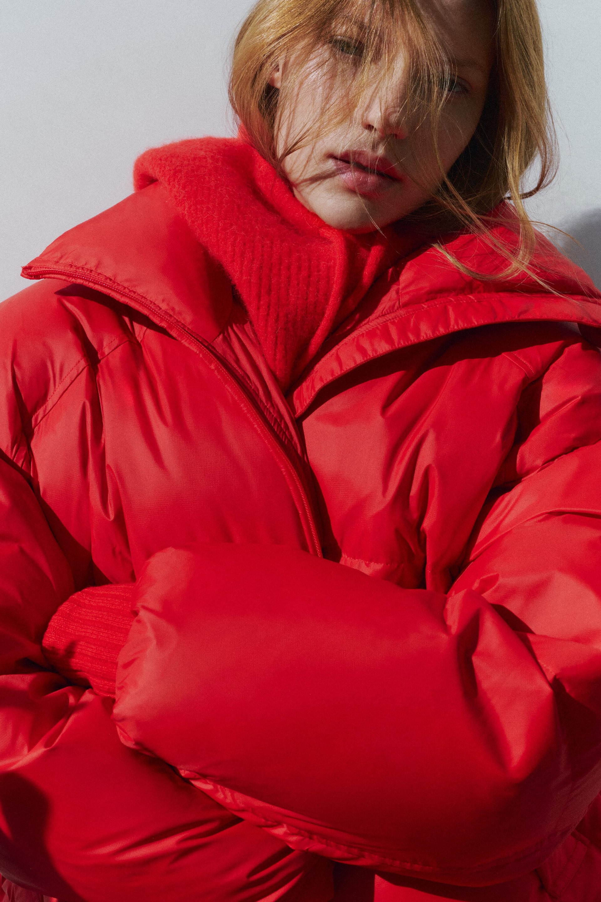 Größeres Bild ansehen: Eine leuchtend rote Steppjacke wird von vorn getragen und zeichnet sich durch ein voluminöses, gestepptes Design mit einem breiten, hohen Kragen und einem Reißverschluss vorne aus. Die Jacke wird über einem farblich passenden, roten, strukturierten Strickkragen getragen, während die Person die Arme verschränkt, um die voluminöse Struktur der Ärmel zu betonen.