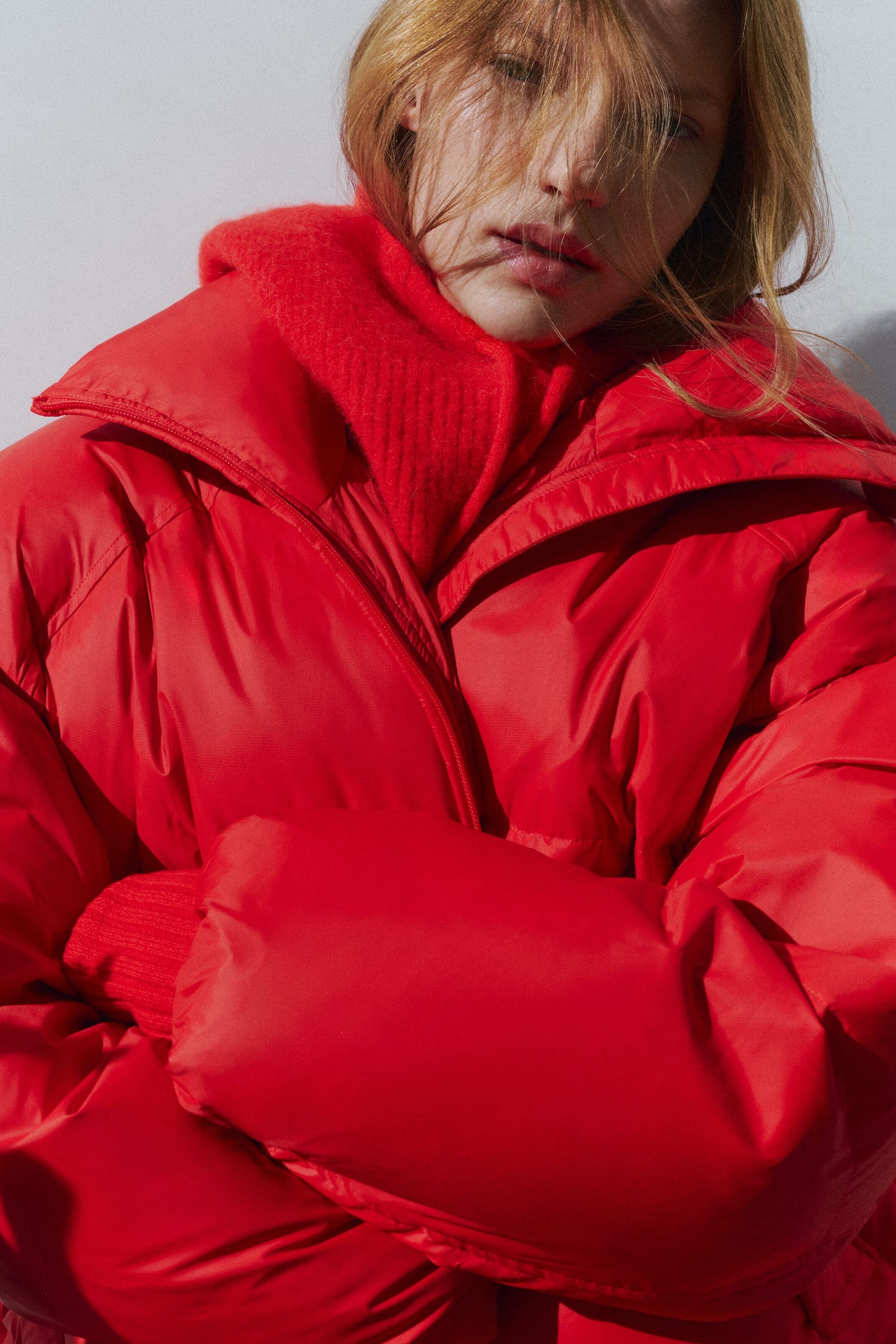 Une doudoune rouge vif est portée de face, avec un design matelassé volumineux, un col montant large et une fermeture zippée sur le devant. La veste est superposée sur un col en maille texturée rouge assorti, tandis que la personne croise les bras pour mettre en valeur la structure bouffante des manches. - 1