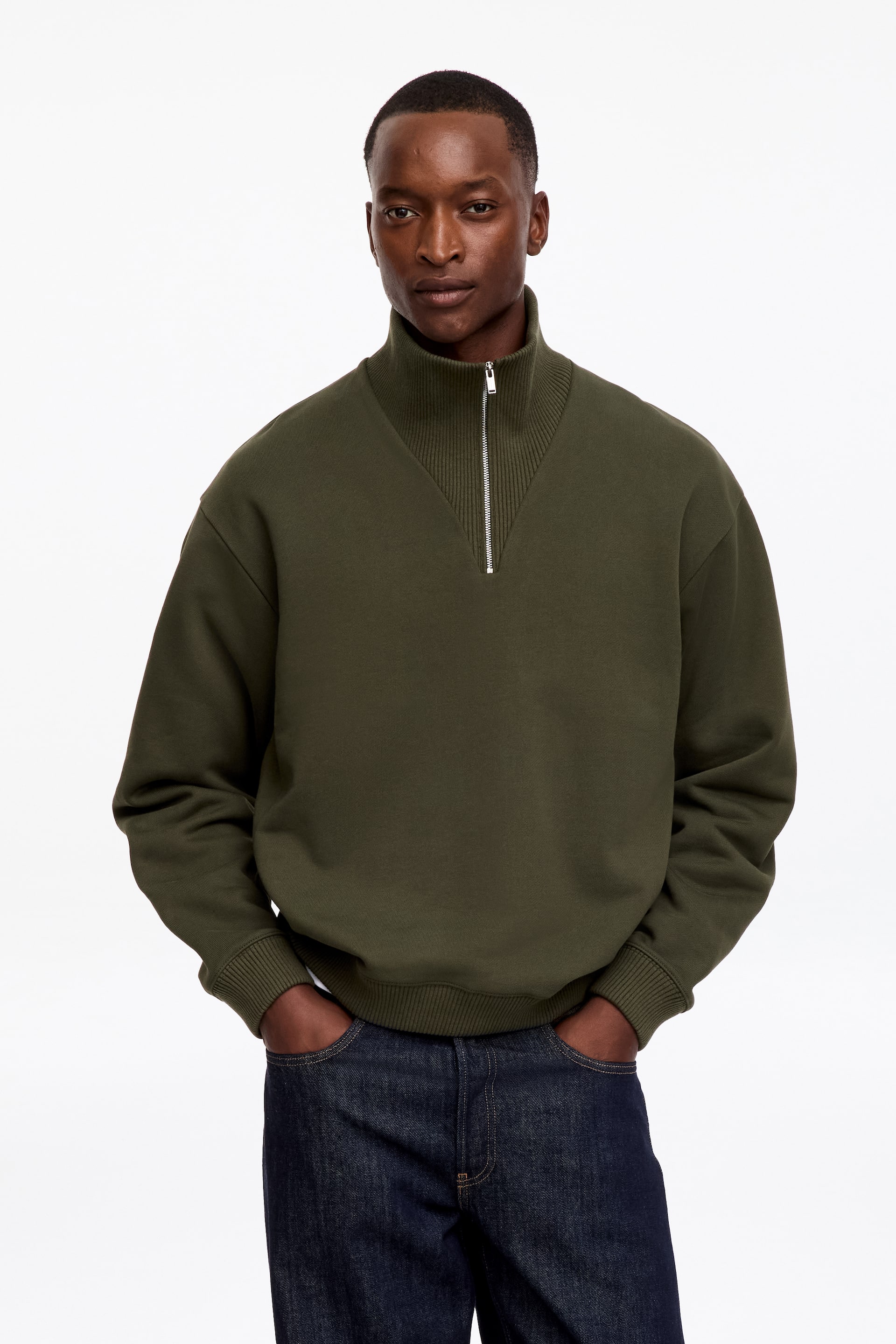Agrandir l'image: Un sweat-shirt vert olive, doté d’un col montant côtelé avec fermeture zippée métallique à demi-zip, de poignets côtelés et d’un ourlet côtelé, est porté par le mannequin. Le sweat-shirt présente une coupe regular.