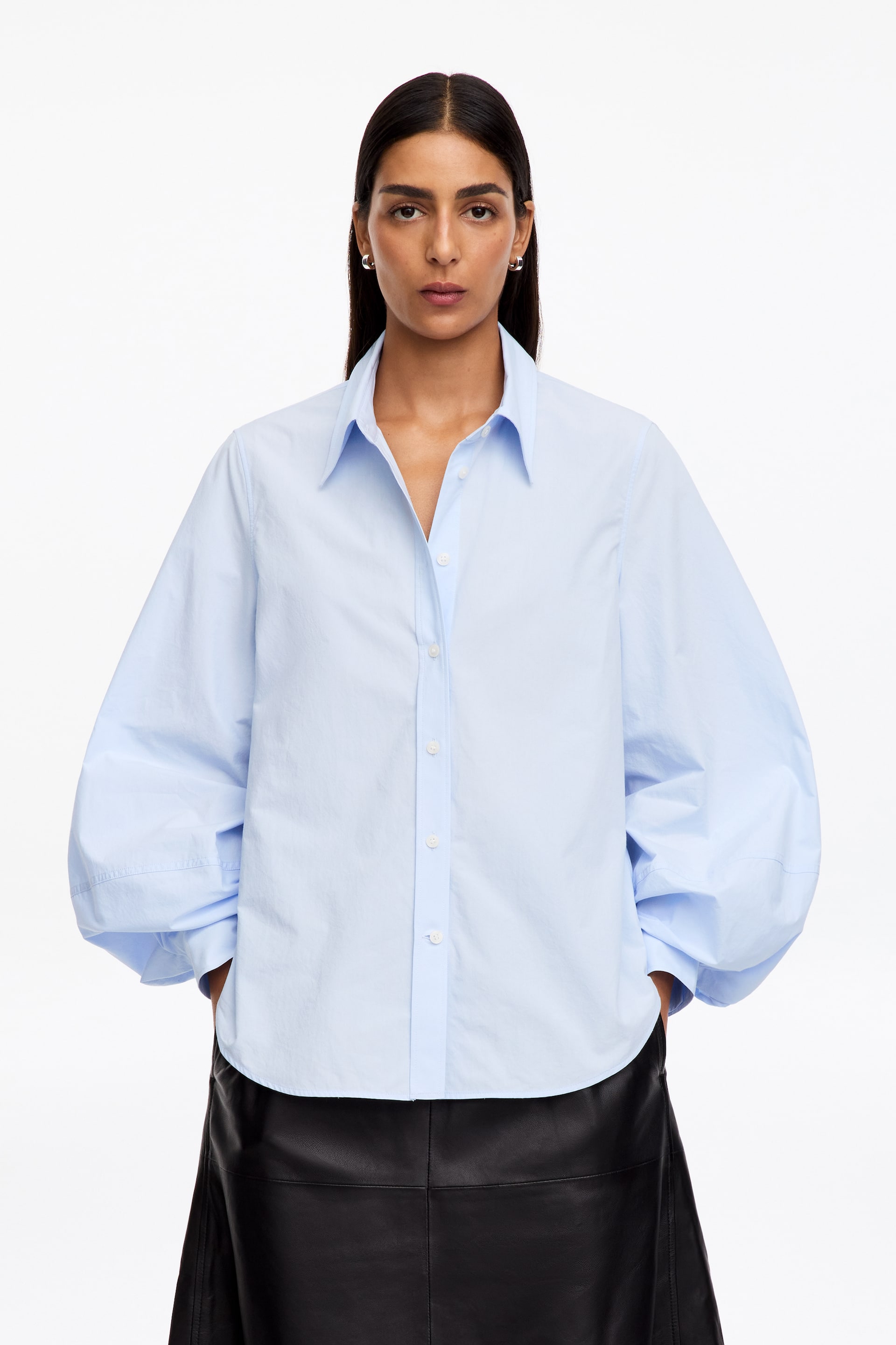 Visualizza l'immagine ingrandita: La modella indossa una camicia azzurro chiaro, caratterizzata da un colletto classico, abbottonatura frontale e maniche lunghe e voluminose. La camicia ha un orlo arrotondato e la modella tiene le mani nelle tasche di una gonna nera.