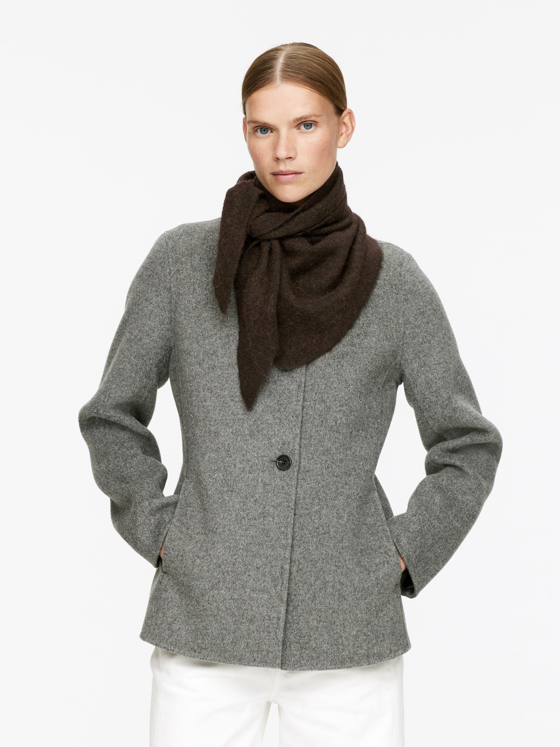 Agrandir l'image: Un manteau gris, à simple boutonnage, de longueur aux hanches, avec une fermeture à un seul bouton foncé et deux poches fendues à l'avant est présenté. Une écharpe en tricot volumineuse et marron foncé est enroulée et nouée autour du cou, superposée au manteau.