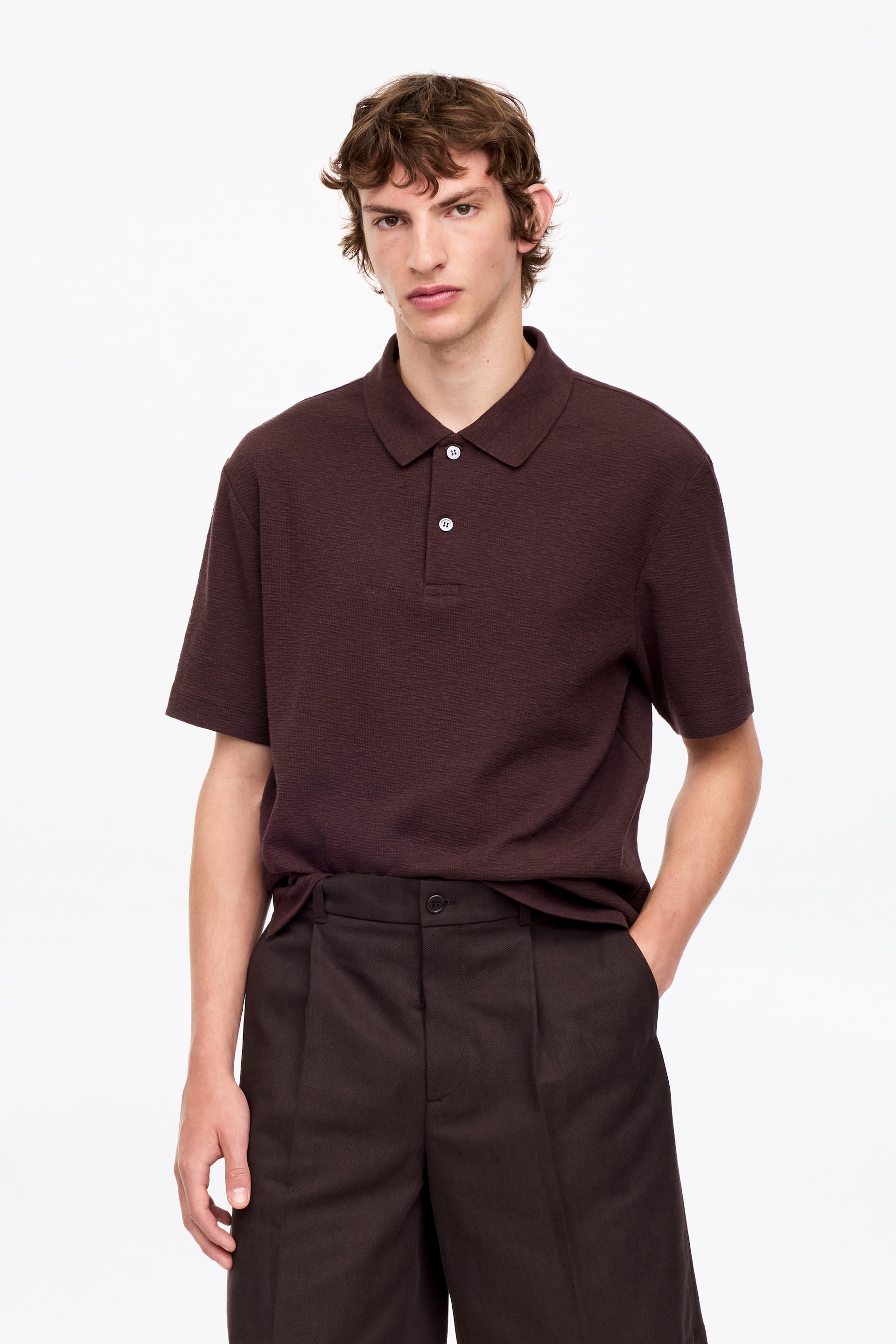 Agrandir l'image: Un polo marron foncé présente un tissu texturé, un col classique et une patte de boutonnage à trois boutons avec deux boutons blancs visibles. Il est doté de manches courtes et d'une coupe décontractée.