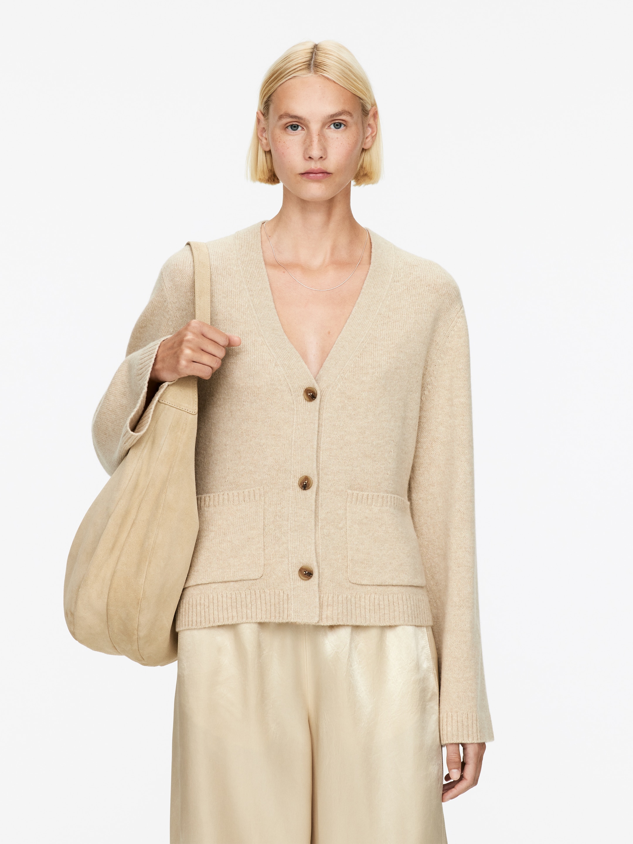 Agrandir l'image: Un cardigan beige clair à col en V est présenté de face, mettant en valeur trois boutons marron au centre et deux grandes poches plaquées sur le bas devant. Le cardigan présente des poignets côtelés et un ourlet côtelé, et est porté par-dessus un pantalon de couleur claire.