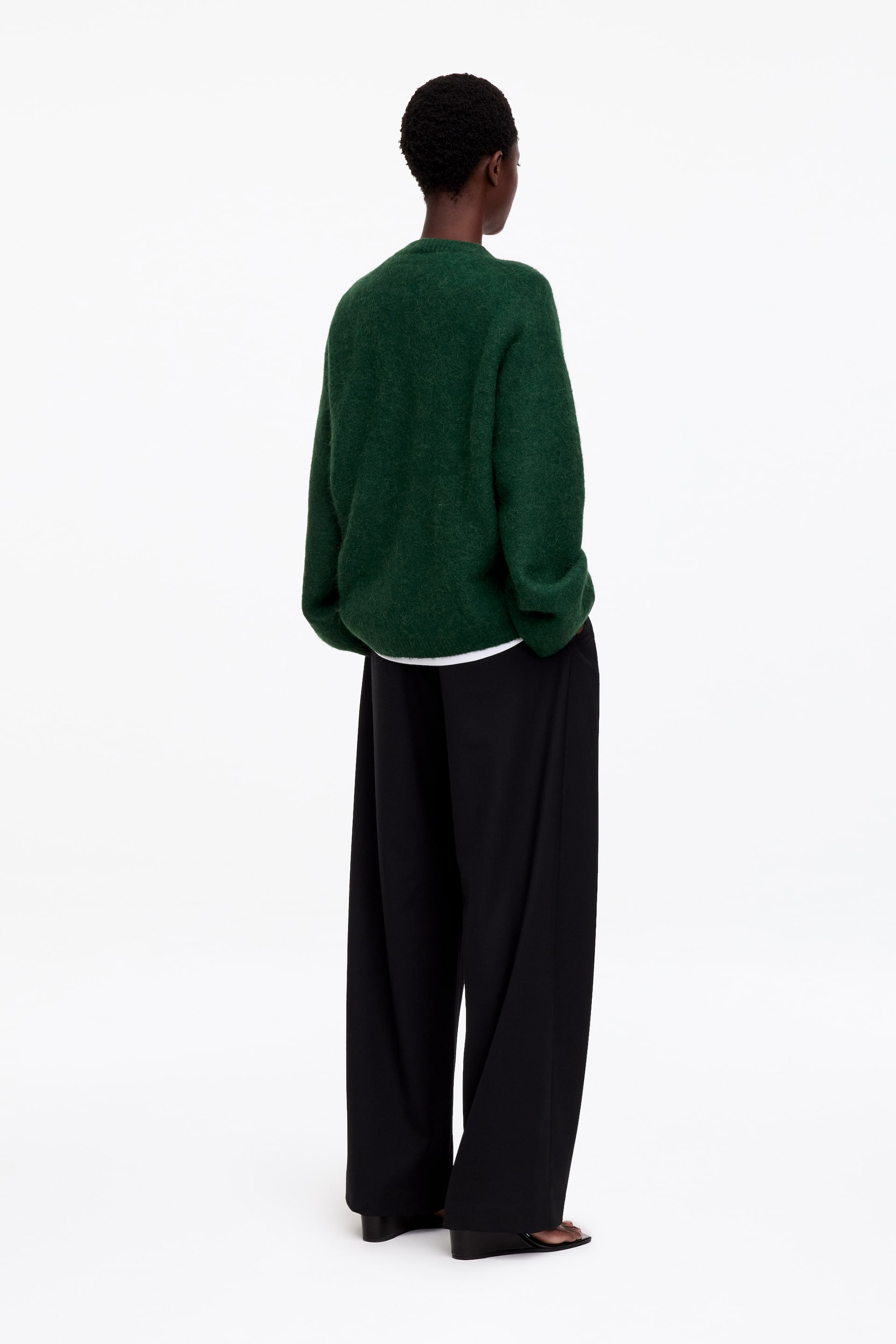 Agrandir l'image: Un pull en maille texturée vert foncé est porté par le mannequin, présenté en vue arrière trois-quarts. Ce pull à coupe décontractée est superposé à un t-shirt blanc et associé à un pantalon noir à jambe large.