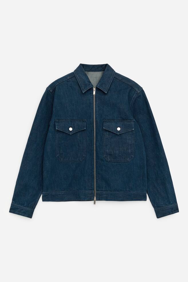 En mörkblå denimjacka visar sin FRAMSIDA, med en fullängds silverdragkedja. Två fickor med lock och silverknappar syns på bröstet, och den är designad med en spetsig krage och långa ärmar. - 1