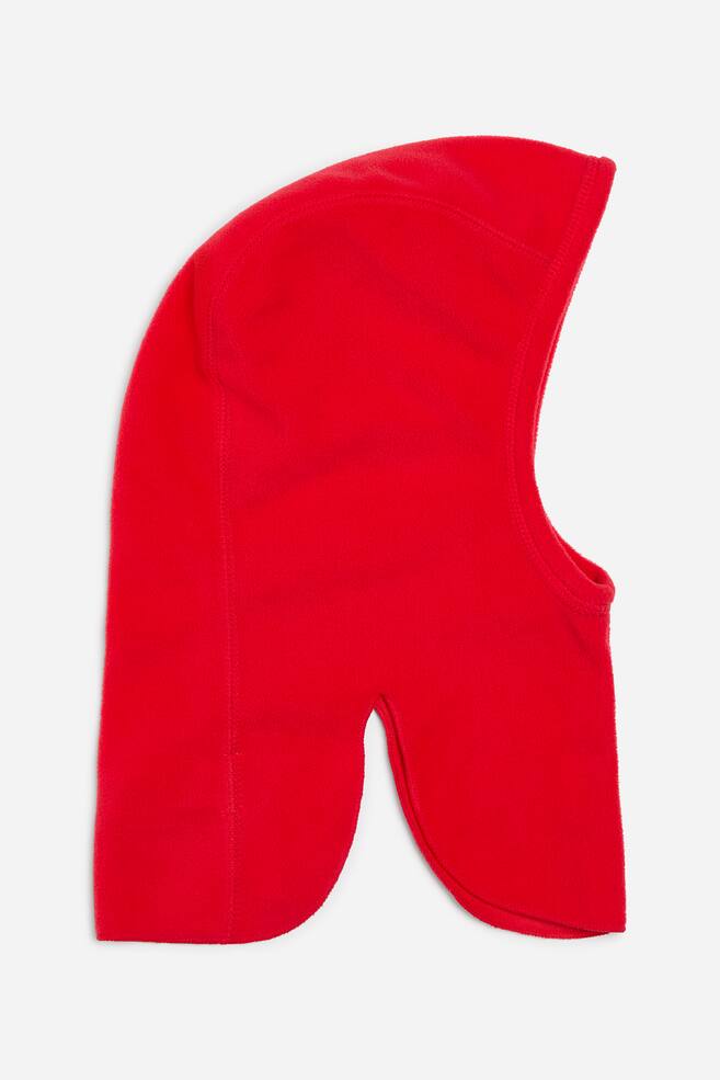 A red fleece hood is shown in side profile, showcasing its soft texture and structured design. The hood features flatlock stitching along its contoured seams, a wide face opening, and high, curved side slits at the hem that create separate front and back panels. - 1