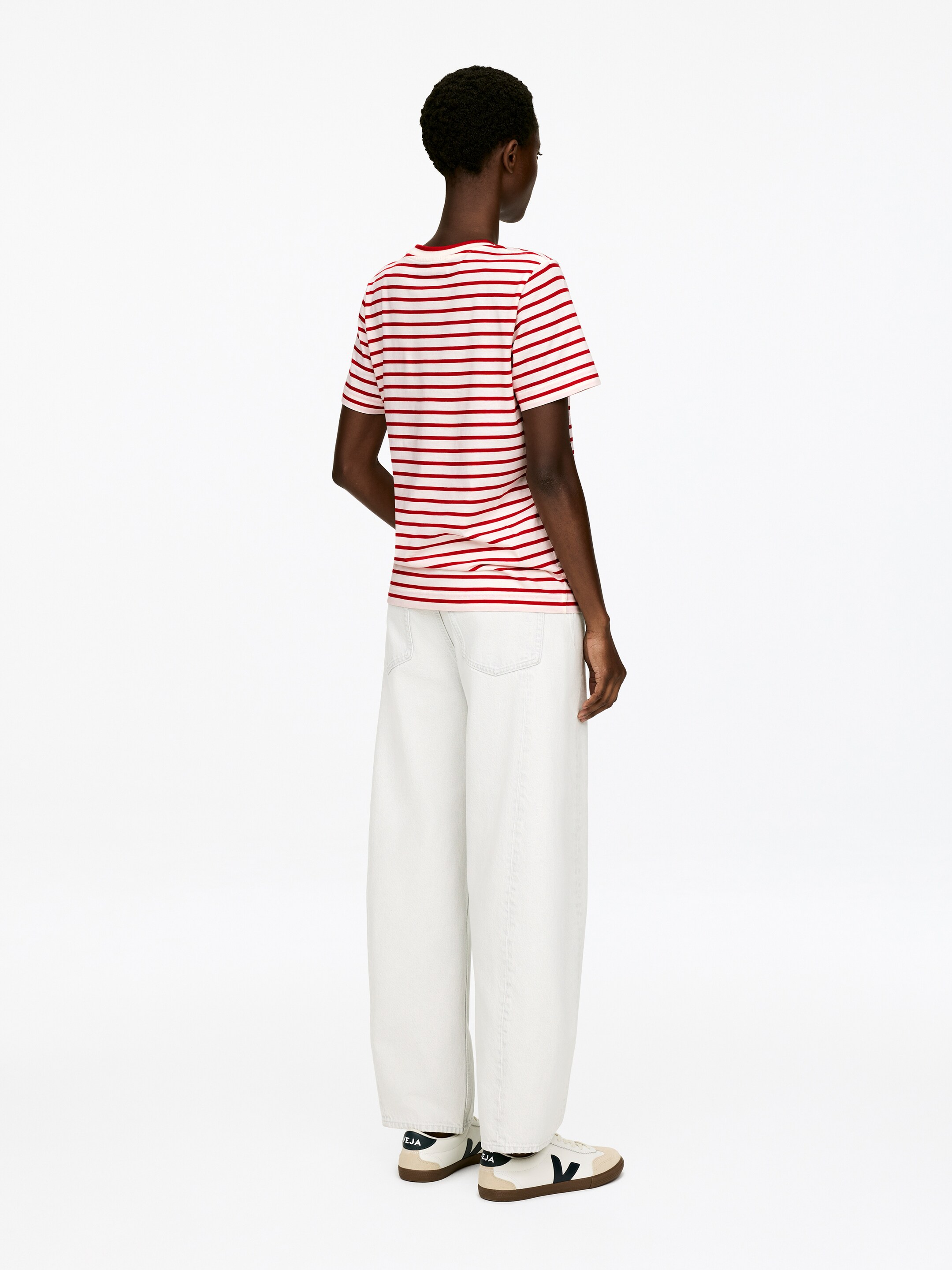 Grotere afbeelding bekijken: Een t-shirt met korte mouwen toont rode en off-white horizontale strepen, met de rug naar de kijker gericht. Het wordt gestyled met lichtcrème, wijde broekspijpen en off-white sneakers met een donker V-logo.