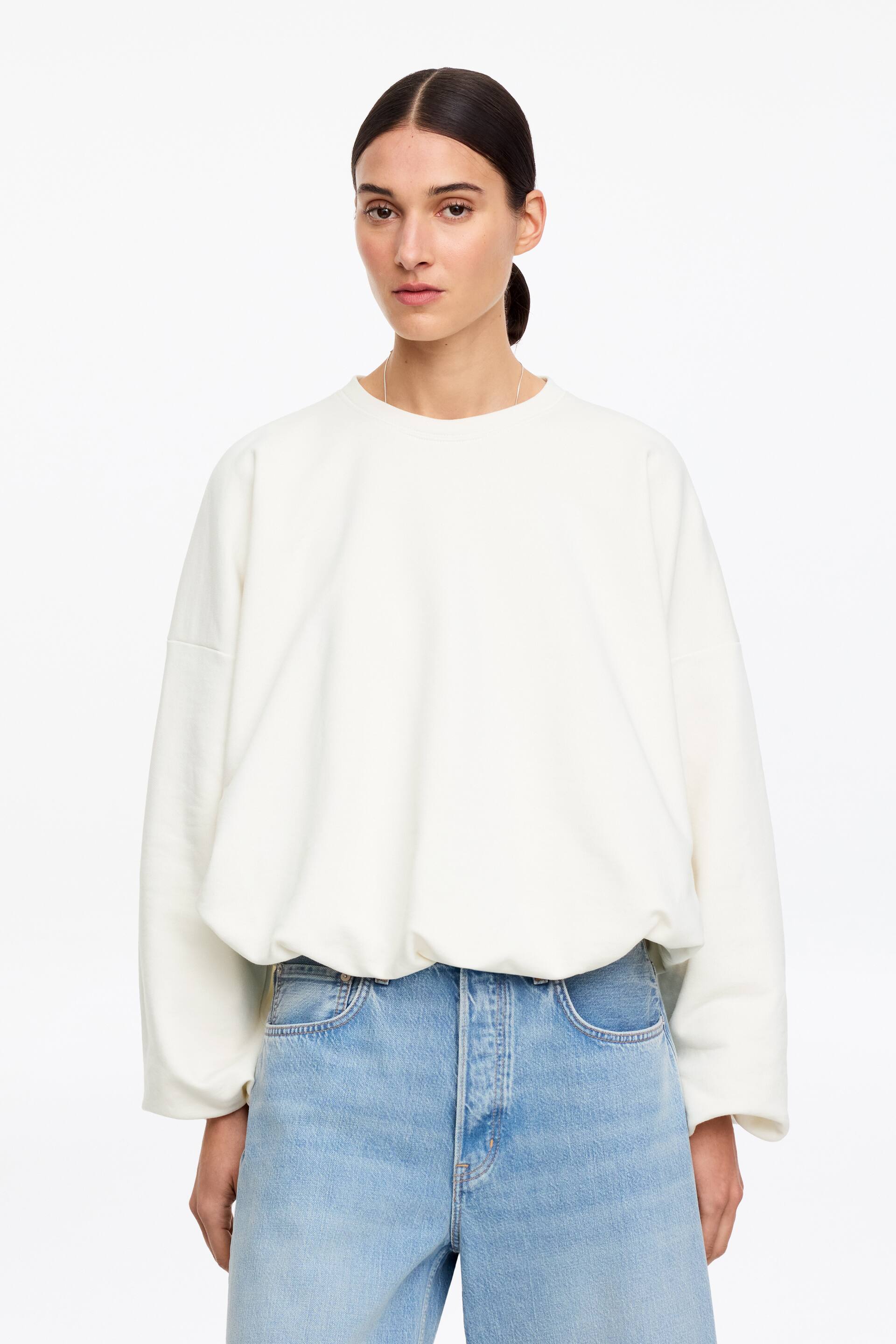 Se større billede: En cremefarvet sweatshirt med rund hals bæres af modellen. Den har voluminøse ballonærmer og en samlet boblekant, der passer over lyse denimjeans.