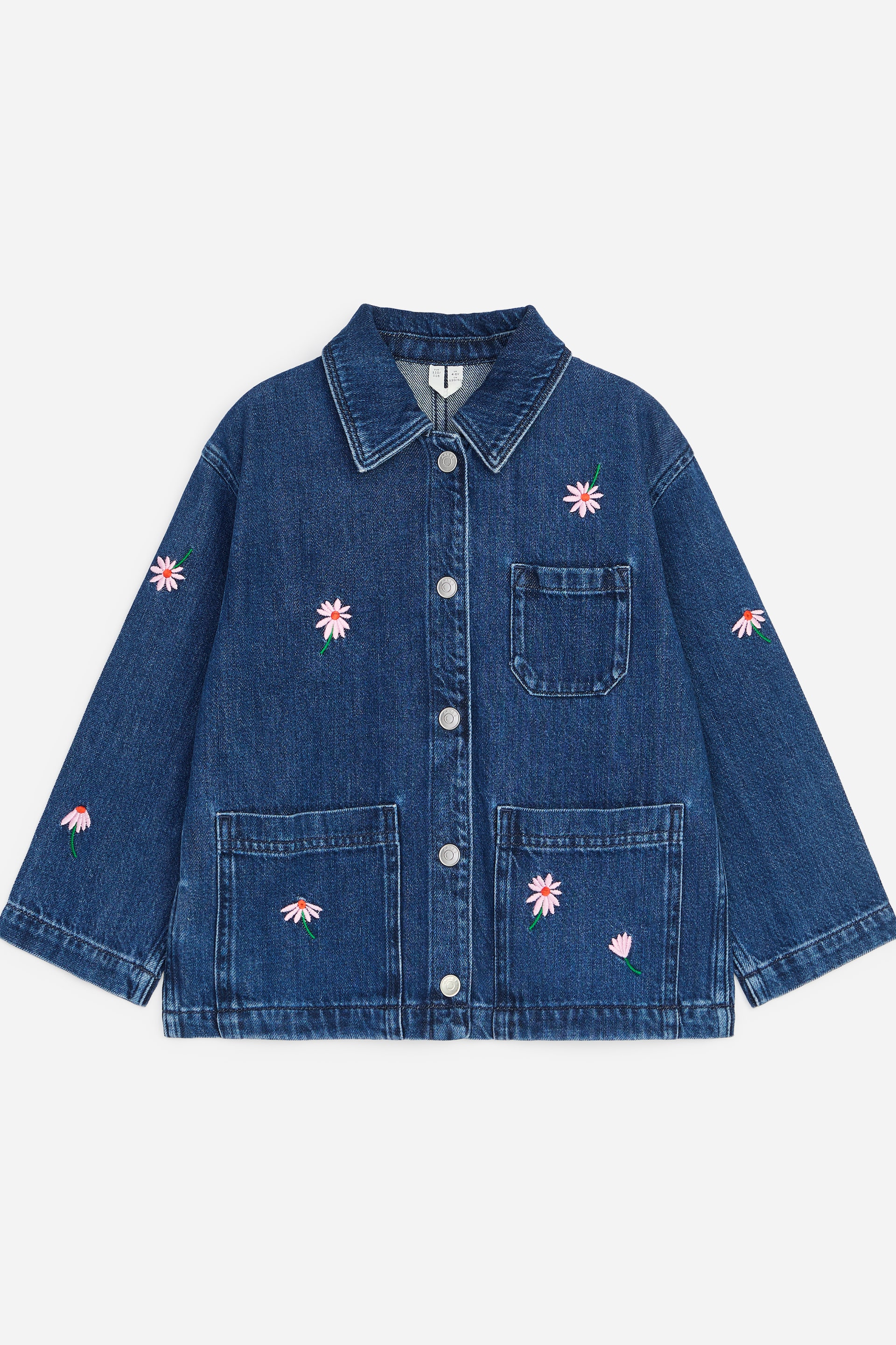 Se større billede: Den blå denimjakke præsenterer sin front, med en spids krave, en fuld knaplukning med sølvfarvede knapper og tre påsyede lommer. Pink og hvid margueritlignende blomster er broderet på forsiden og de lange ærmer.
