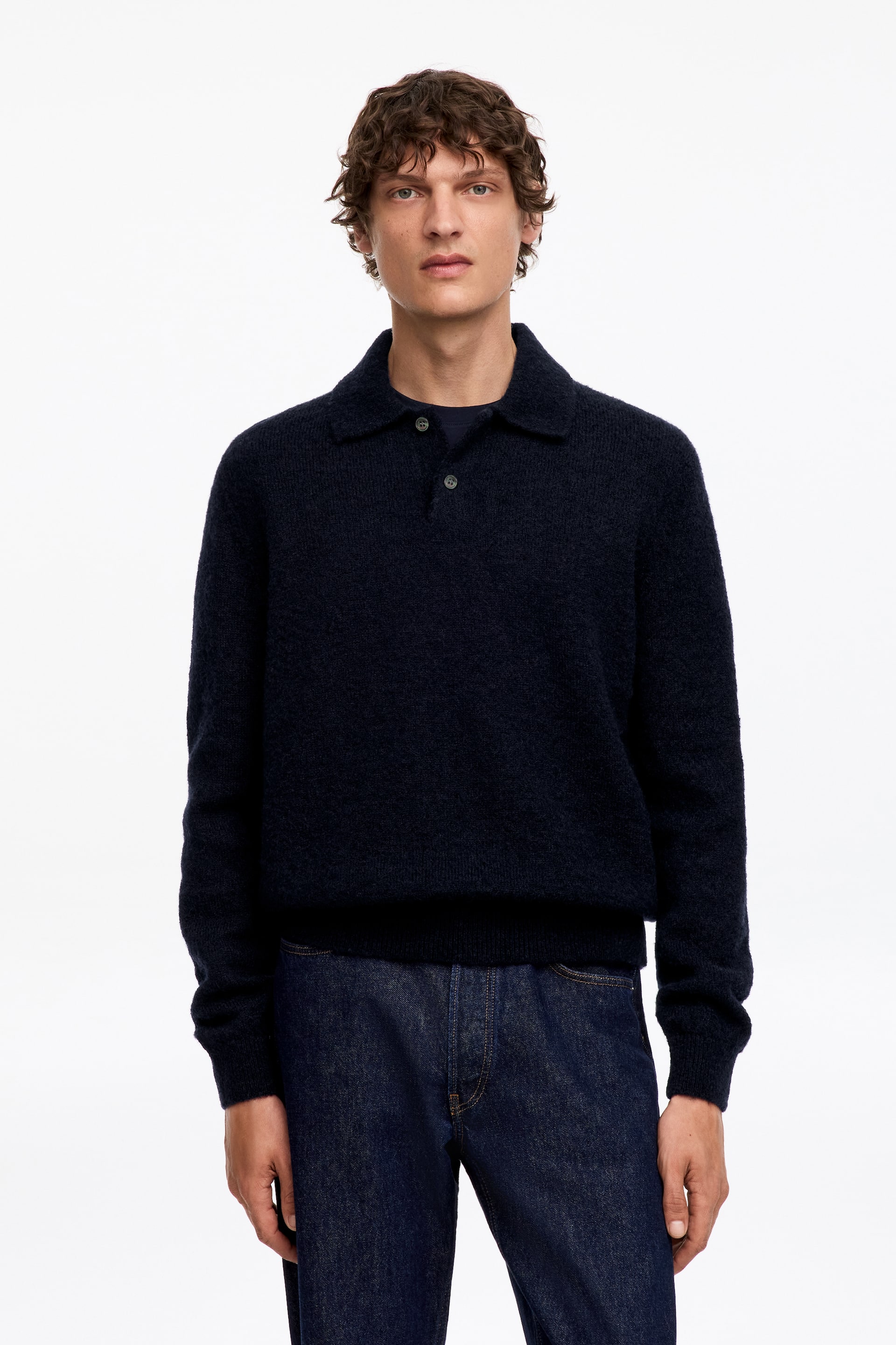 Agrandir l'image: Un polo en maille bleu foncé en mélange de laine, doté d'un col et d'une patte de boutonnage partiellement déboutonnée révélant un t-shirt plus foncé en dessous, est porté par le mannequin avec un jean bleu foncé.