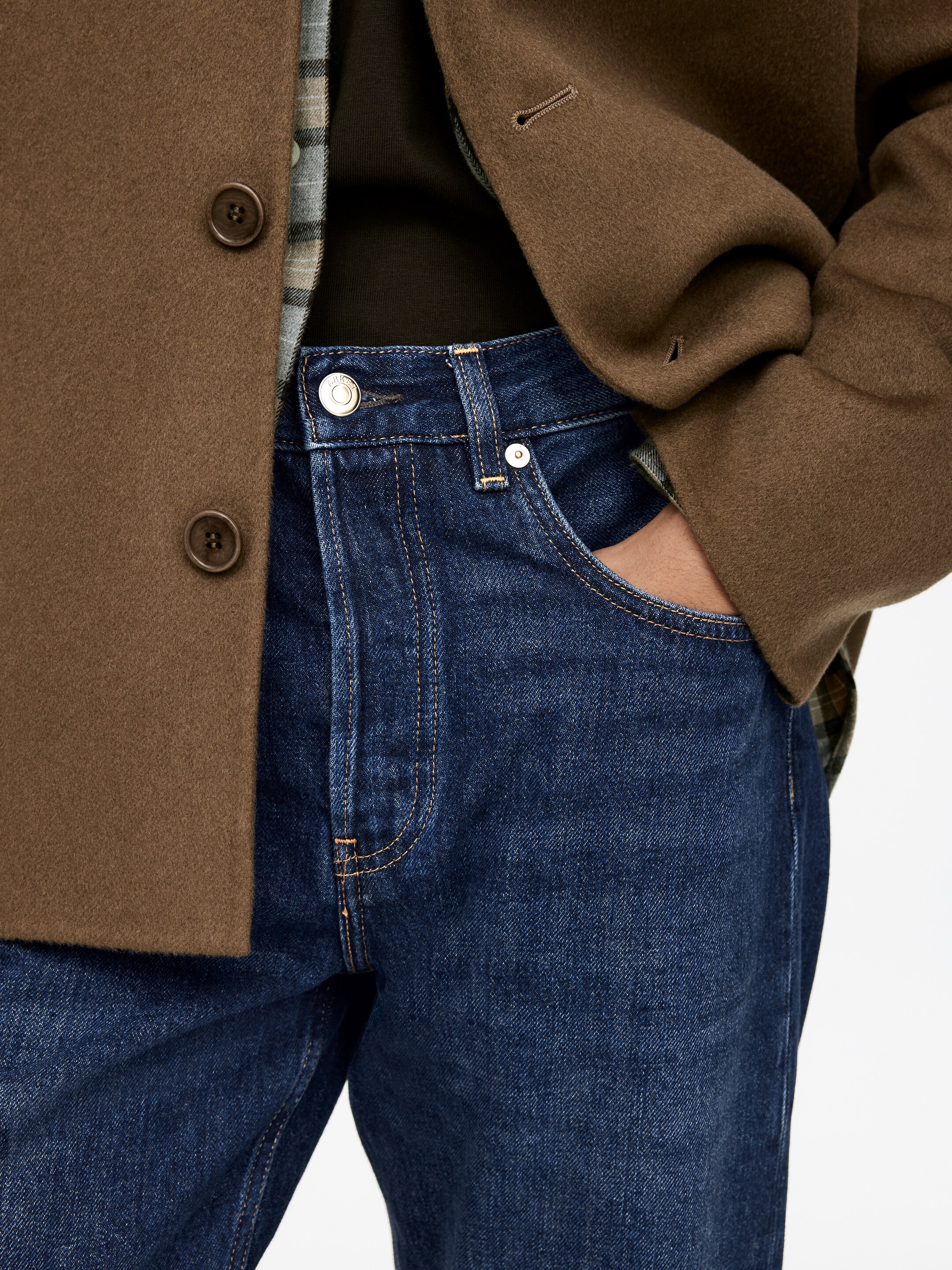 Visa större bild: Mörkblå jeans presenteras, med orange toppsömmar, metallnitar och en metallknapp i midjan, med en hand vilande i den främre högra fickan. En brun ullblandad kappa, med bruna knappar och ett knapphål vid manschetten, är lagrad över en mörkgrön topp och en grönrutig skjorta.