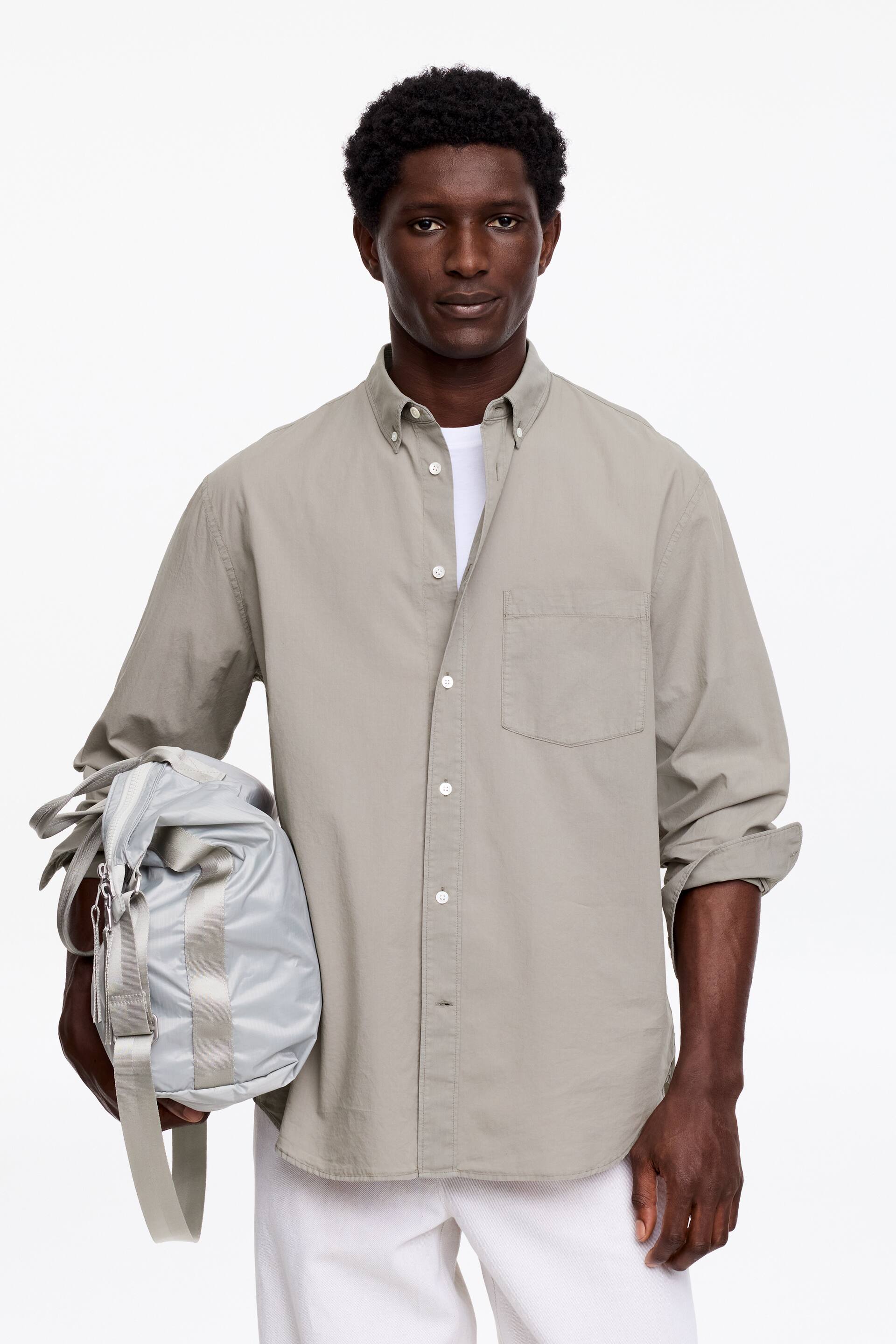 Näytä suurempi kuva: Vaaleanharmaa nappipaita esittäytyy edestäpäin, ja siinä on nappikaulus, täysi nappilista valkoisilla napeilla, yksi rintatasku vasemmalla puolella ja pitkät hihat käärittyinä kyynärvarsille, paljastaen valkoisen t-paidan alla.