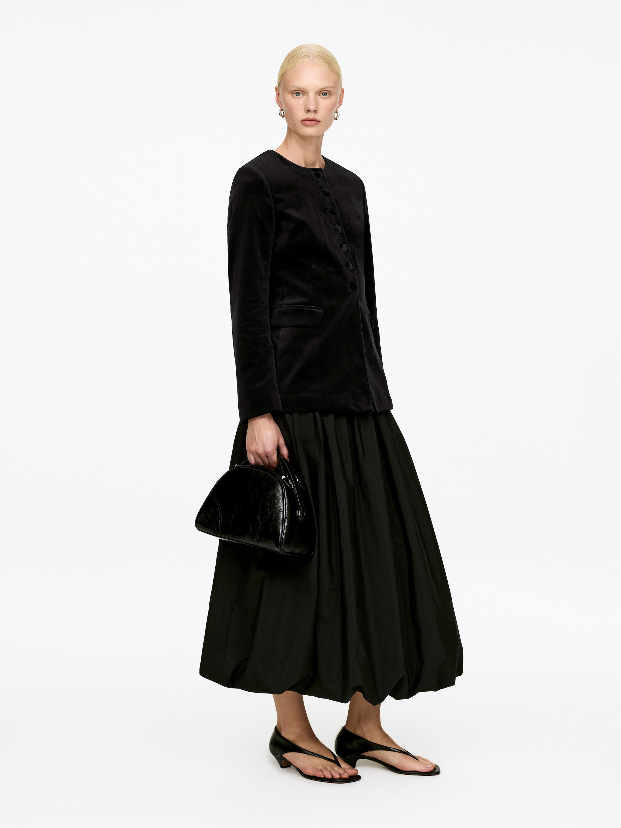 Ver imagen más grande: El conjunto negro se presenta en un ángulo ligeramente inclinado, con una chaqueta de terciopelo negro sin cuello con una tapeta de botones y dos bolsillos con solapa, y una falda midi negra voluminosa con un dobladillo de burbuja fruncido. Un bolso de mano negro estructurado con un asa superior y sandalias negras de tacón bajo con punta tipo tanga complementan el look.