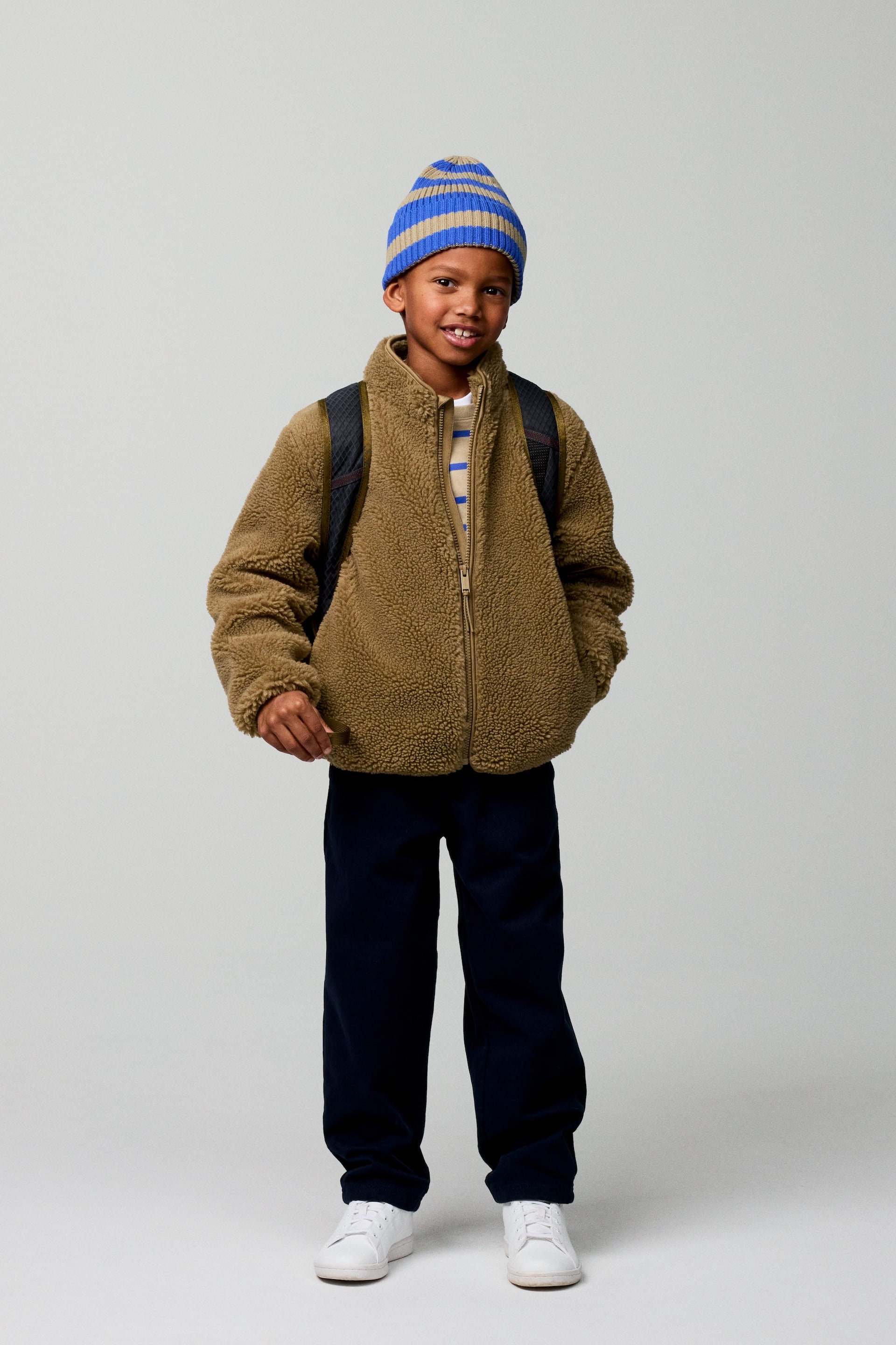 Vis større bilde: En olivengrønn sherpa-jakke med full glidelås og høy krage presenteres forfra, lagd over en lys, horisontalt stripet undertrøye og mørkeblå bukser med rette ben. En blå og beige horisontalt stripet strikket lue bæres sammen med hvite joggesko, og en mørk ryggsekk bæres over jakken.