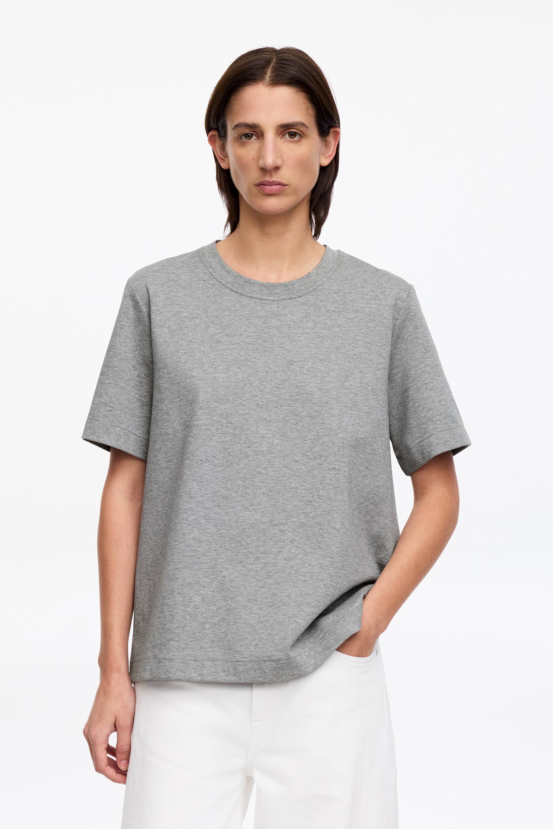 Zobraziť väčší obrázok: Modelka má na sebe sivé tričko s okrúhlym výstrihom, ktoré má krátke, jemne prispôsobené rukávy a rovný lem. Tričko má bežný strih, keď modelka stojí, s jednou rukou opretou o bok a druhou rukou siahajúcou do vrecka bielych nohavíc.