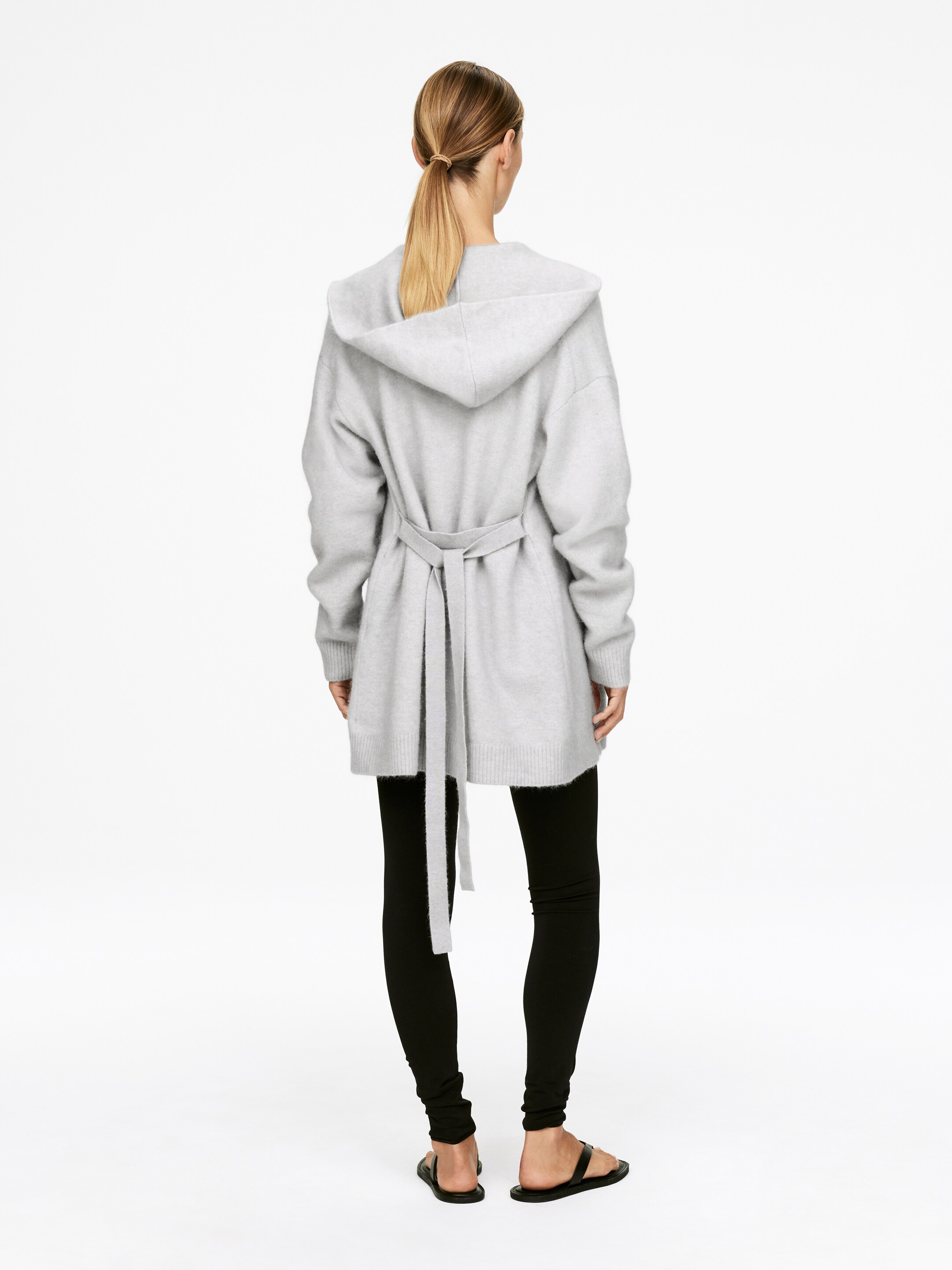 Vis større bilde: En lys grå hettegenser-omslag cardigan vises fra baksiden. Den har en avslappet passform med et belte i samme stoff som strammer inn i midjen, og strekker seg til midt på låret over svarte leggings.