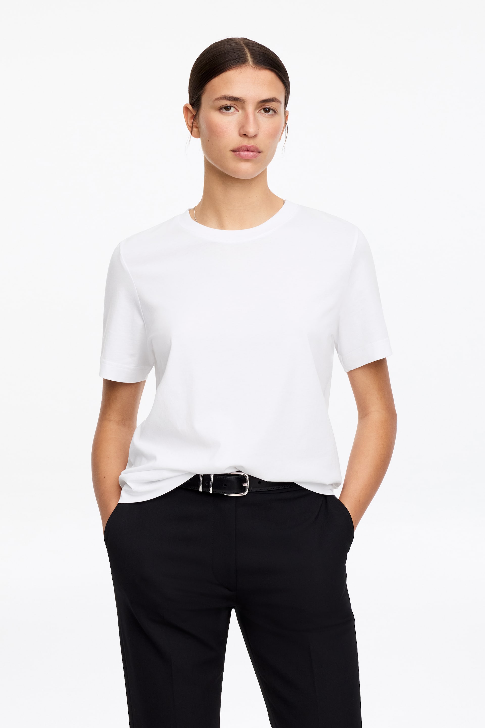 Visa större bild: En vit t-shirt visas med rund hals och korta ärmar i en framåtvänd vy. Den har ett slätt tyg och bärs instoppad i svarta byxor.