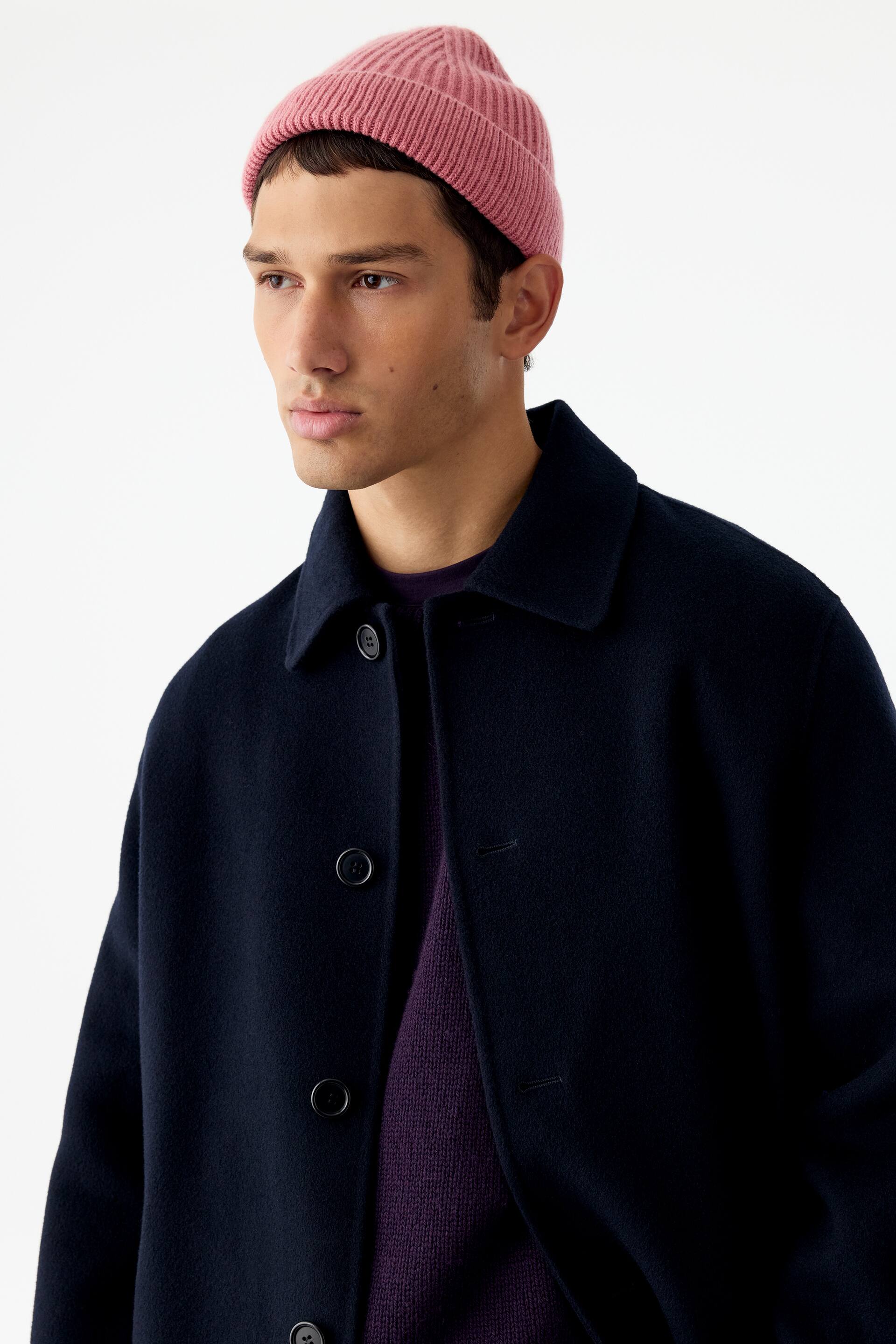View larger image: A pink, ribbed-knit beanie with a folded brim is being worn by the model. The beanie is paired with a dark navy buttoned coat and a purple knitted jumper.