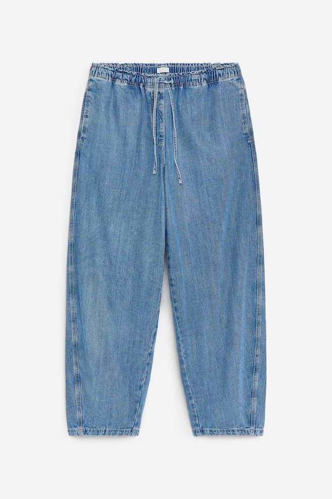 DEVANT. Un jean en denim bleu clair présente une taille élastiquée et froncée avec un cordon de serrage visible, offrant une coupe décontractée à jambes larges se terminant par un ourlet simple. - 1