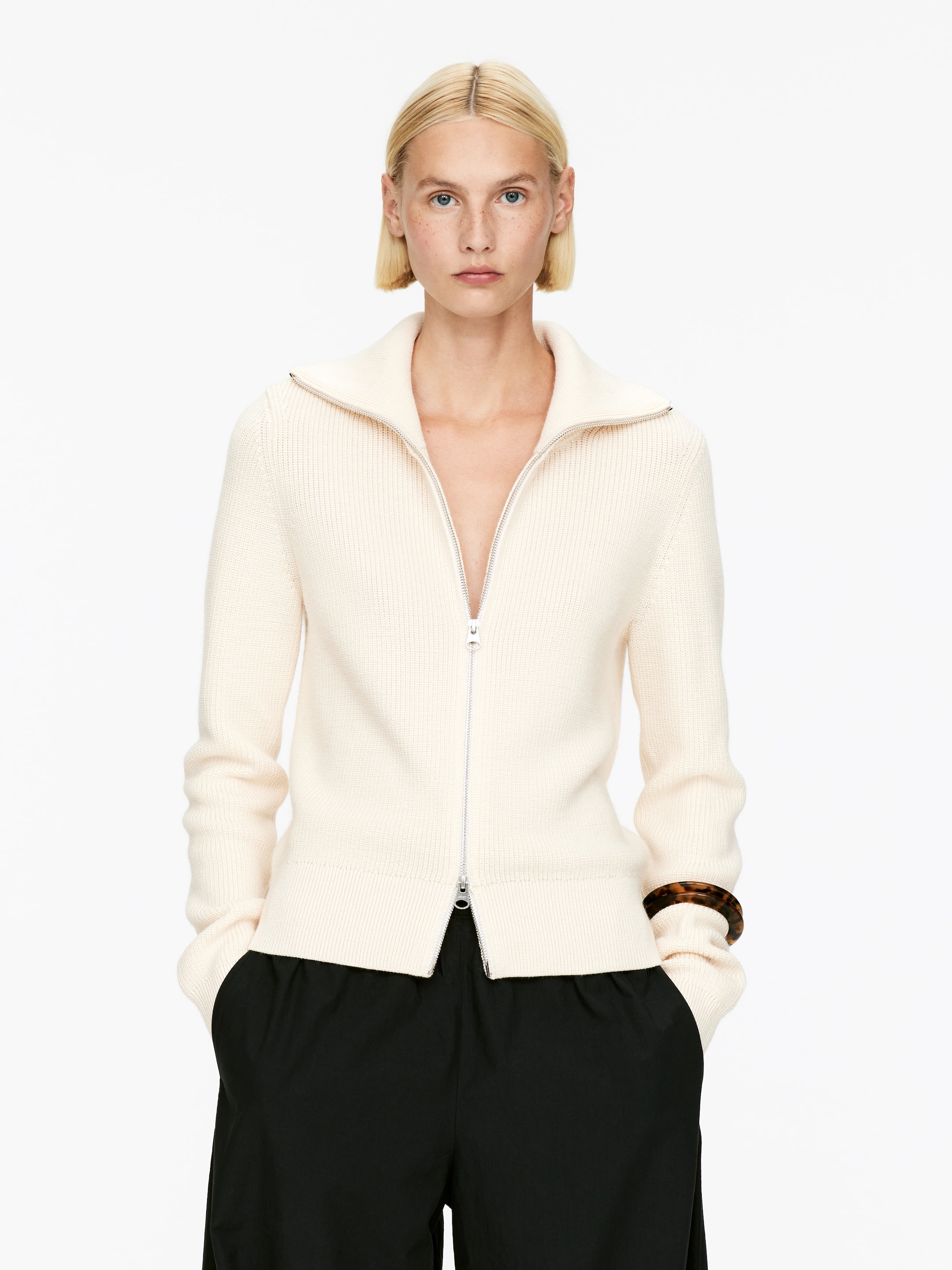 Agrandir l'image: Un pull zippé de couleur crème est présenté de face, mettant en avant sa structure et son ajustement. Le pull est doté d'un col montant, d'une fermeture éclair argentée à double sens sur toute la longueur, et de détails en maille côtelée visibles sur le col, les poignets et l'ourlet.