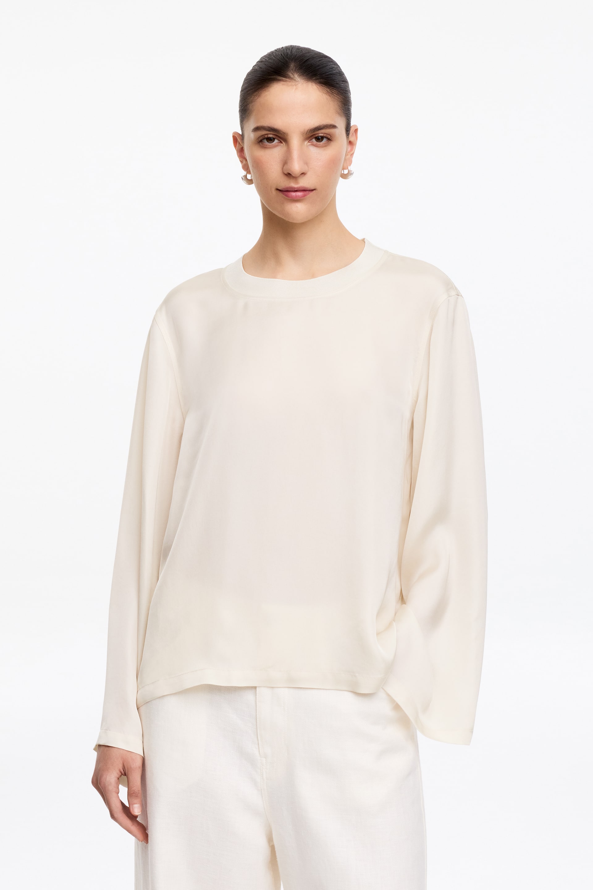 View larger image: The cream long-sleeved top is facing forward, featuring a crew neck and loose, flowing sleeves ending in wide openings, and is loosely resting over white trousers.