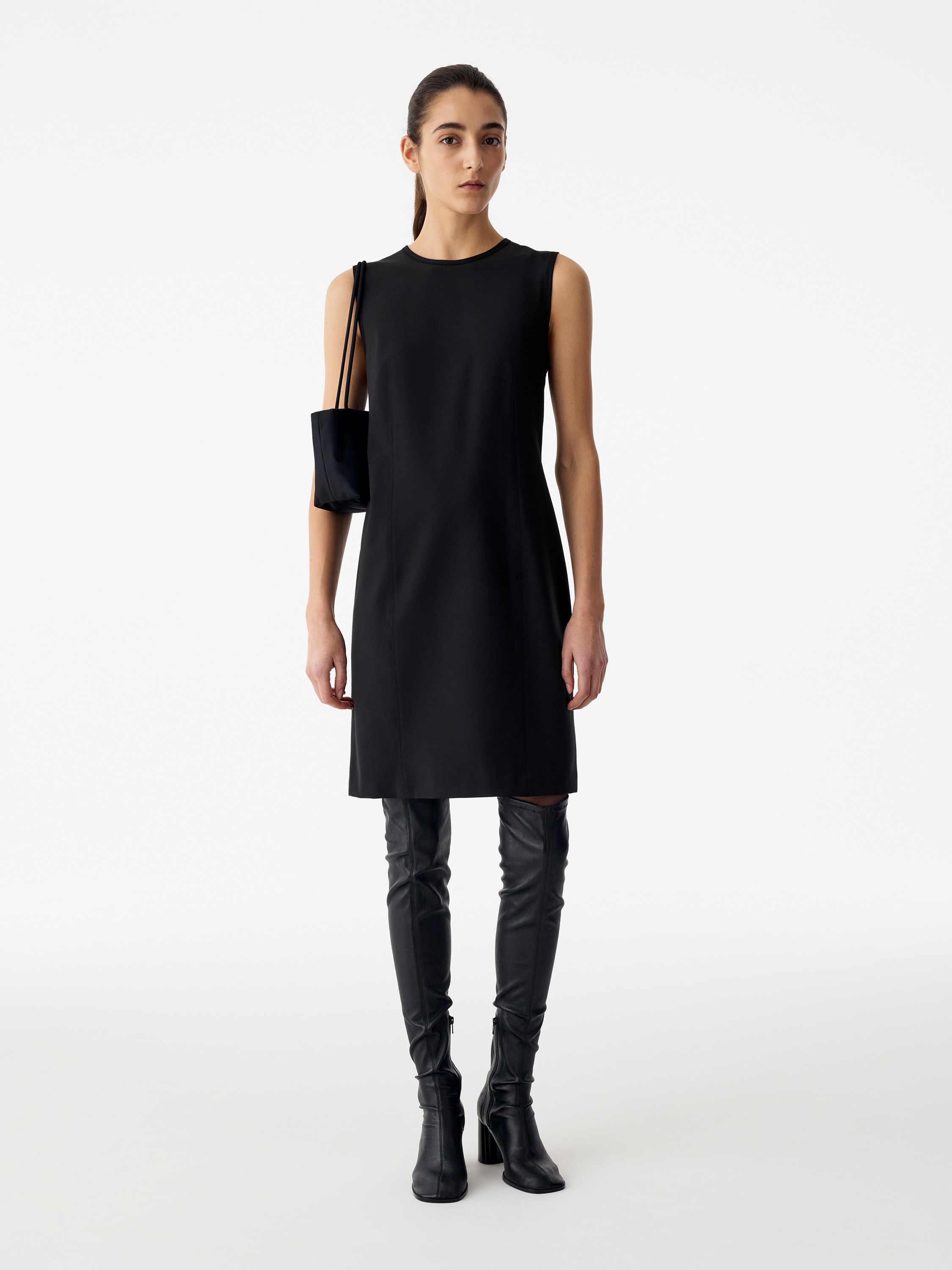 Um vestido preto sem mangas, estilo shift, visto de frente, com decote redondo à frente e costuras verticais estruturadas que descem pela frente. O vestido apresenta uma silhueta minimalista, ligeiramente em A, terminando num rebordo direito.
