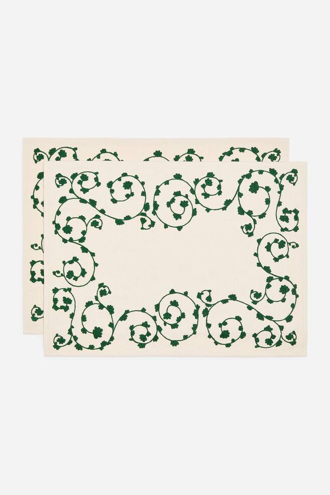 TvÃ¥ rektangulÃ¤ra benvita bordstabletter ligger plant i en framÃ¥triktad vy, dÃ¤r den ena Ã¤r lÃ¤tt placerad bakom den andra. Varje bordstablett visar en grÃ¶n broderad kant av slingrande rankor och blad runt en enfÃ¤rgad rektangulÃ¤r mittdel. - 1