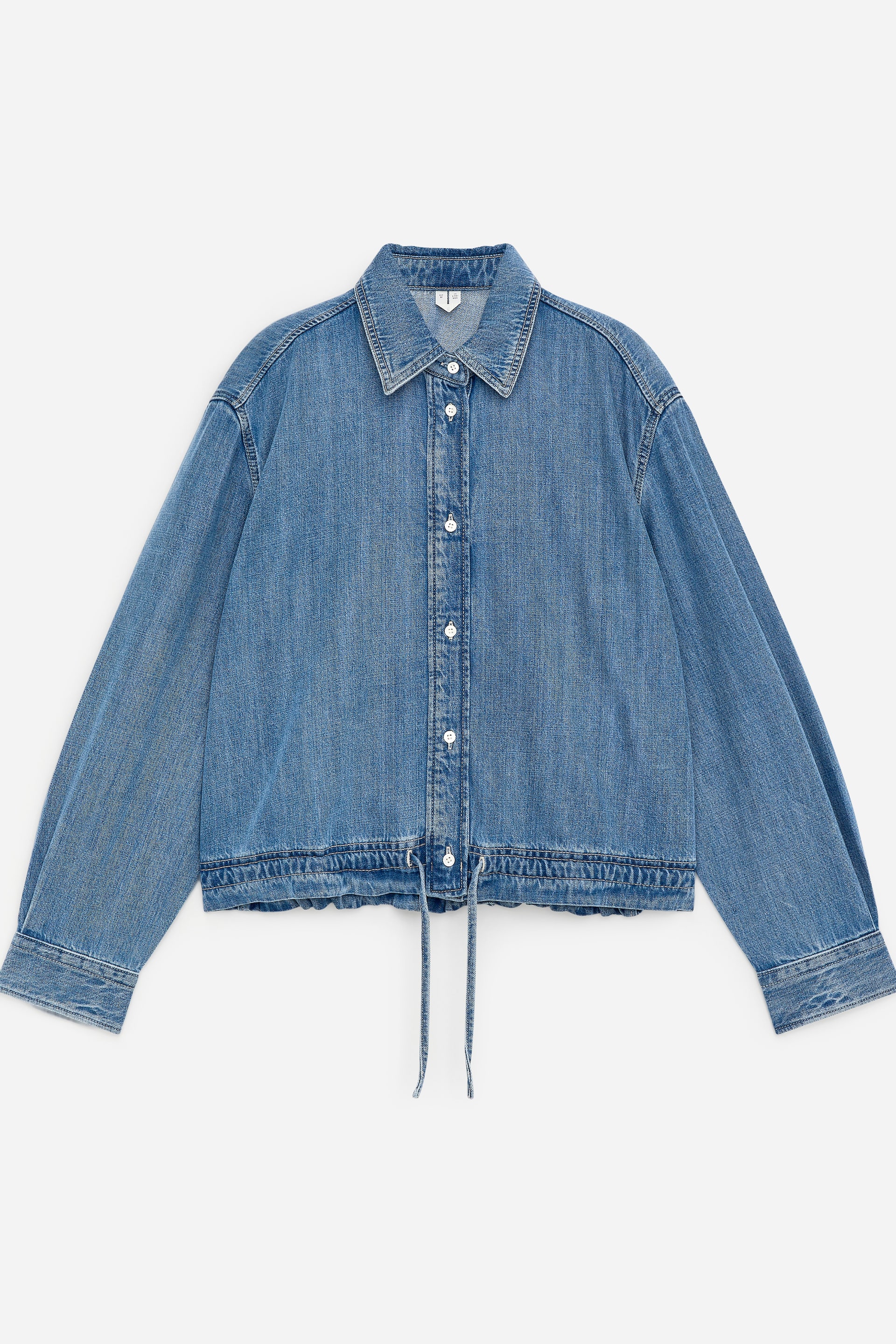 Vis større bilde: FRONT, en lyseblå denimjakke med spiss krage, knappestolpe med hvite knapper, lange ermer med knappede mansjetter, og en innsnevret snor nederst med to bånd som henger sentralt.