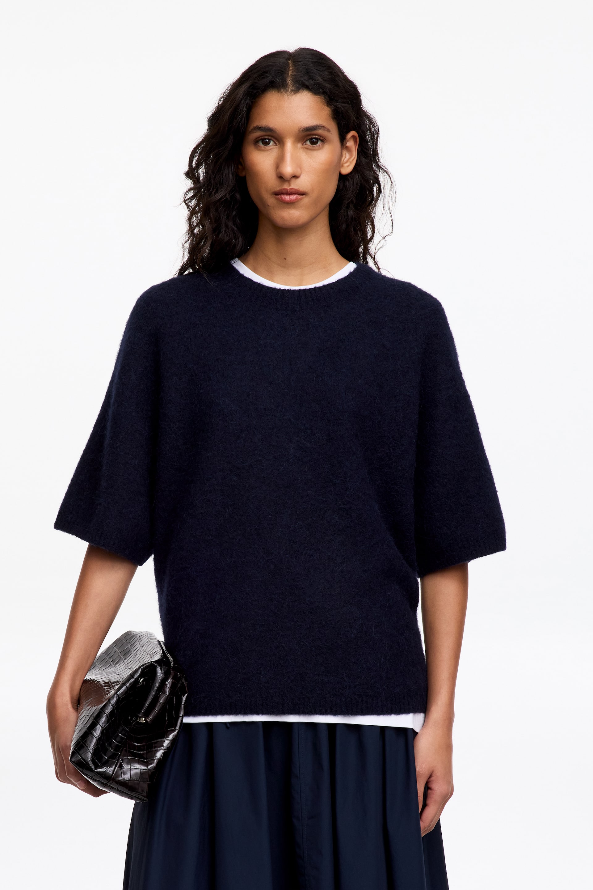 Visualizza l'immagine ingrandita: La modella indossa un maglione a maniche corte in maglia morbida color blu navy scuro, con girocollo e ampie maniche a tre quarti. Il maglione è abbinato a una T-shirt bianca, a una gonna blu scuro, ed è completato da una pochette nera effetto coccodrillo tenuta sotto un braccio.