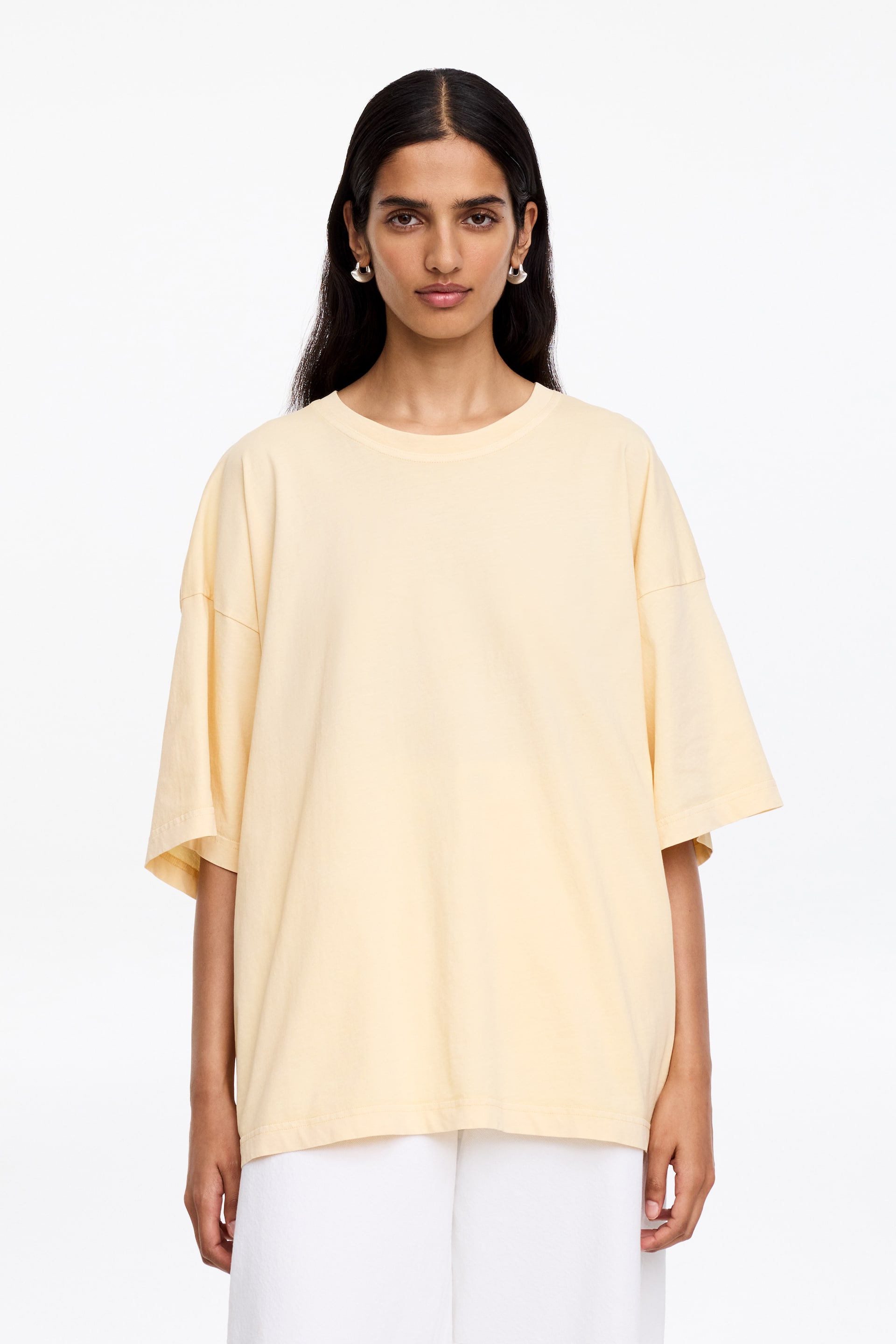 Se større billede: En bleg gul oversized T-shirt bæres af modellen, med en ribbet rund hals, sænkede skuldre og lidt længere korte ærmer. T-shirten falder naturligt over hvide bukser og fremhæver dens afslappede pasform.
