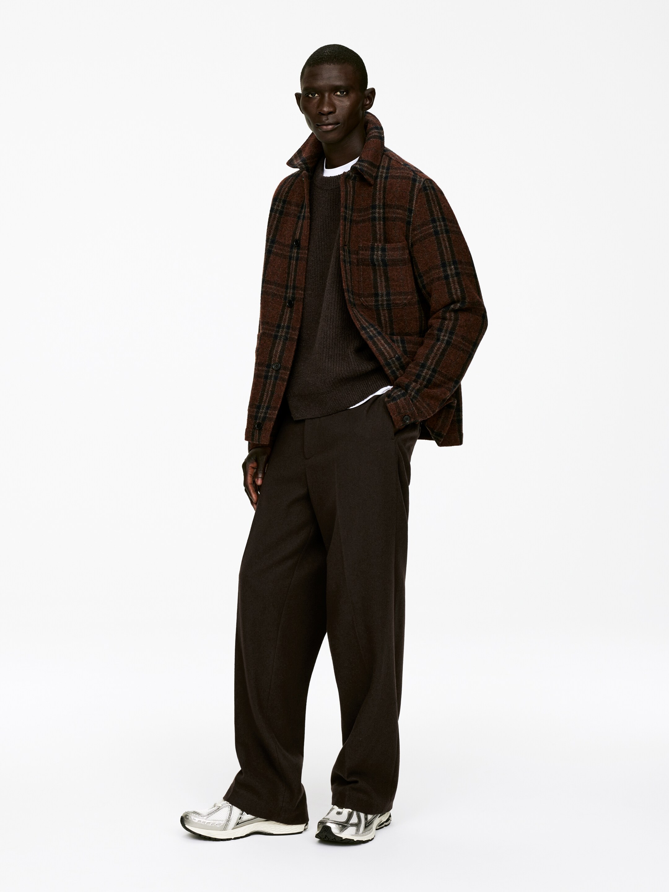 Größeres Bild ansehen: Ein geschichtetes Outfit wird an einer Person präsentiert, die leicht nach rechts geneigt steht. Ein braunes kariertes Overshirt wird offen über einem dunkelbraunen Rundhalspullover mit sichtbaren gerippten Bündchen getragen, das über einer weißen Basisschicht geschichtet ist, während weit geschnittene dunkelbraune Hosen und silberne und schwarze Sportschuhe das Ensemble vervollständigen.