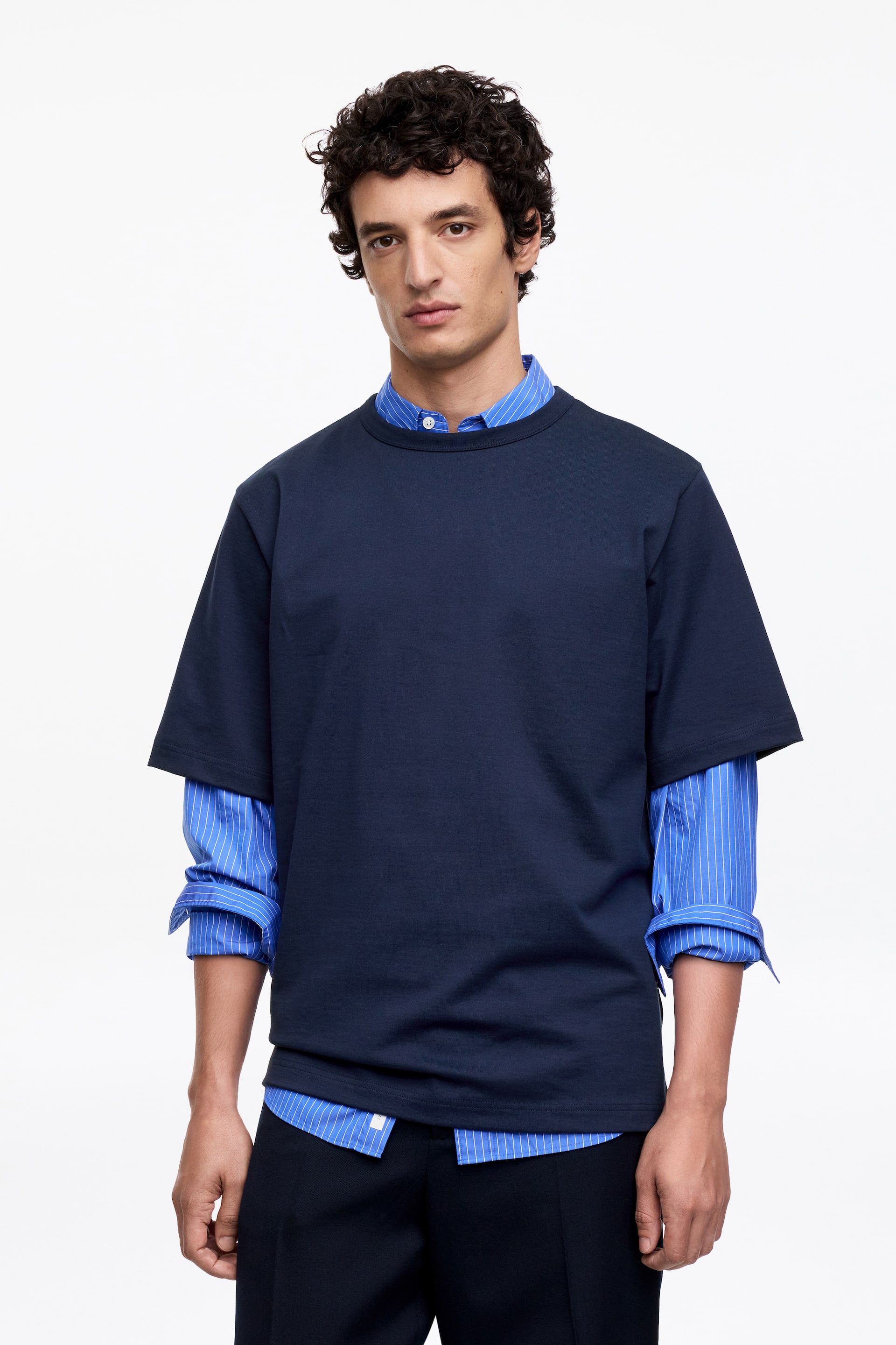 Visa större bild: En marinblå kortärmad t-shirt presenteras, med rund hals och en avslappnad passform. Den är lagrad över en blå- och vitrandig skjorta med krage, där t-shirtens korta ärmar sträcker sig över underplaggets upprullade ärmar.