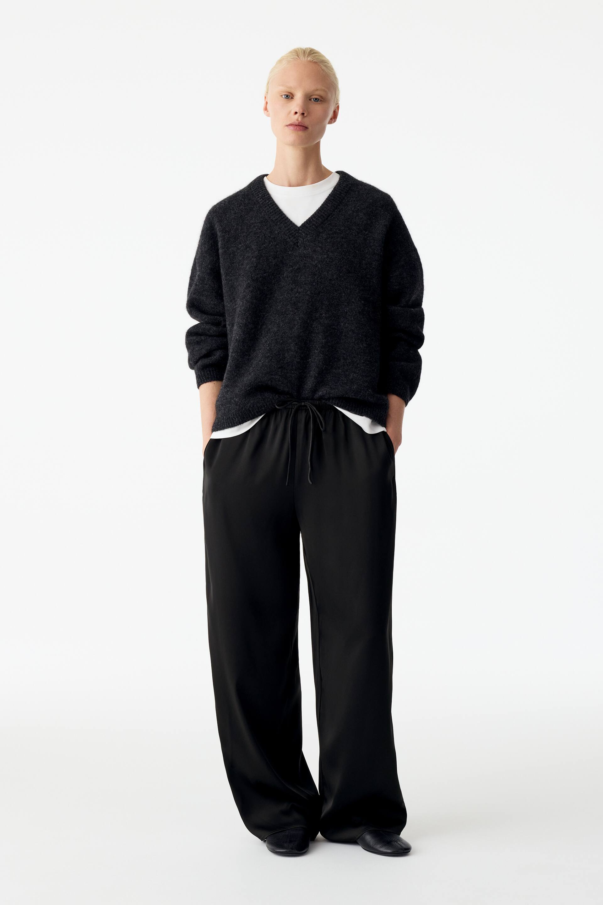 Visualizza immagine più grande: Pantaloni satinati neri dal taglio rilassato, caratterizzati da una silhouette fluida e da una vita con coulisse, indossati dalla modella, che è in piedi con le mani inserite nelle tasche laterali. I pantaloni sono abbinati a un maglione in maglia a V grigio scuro, sovrapposto a una T-shirt bianca.