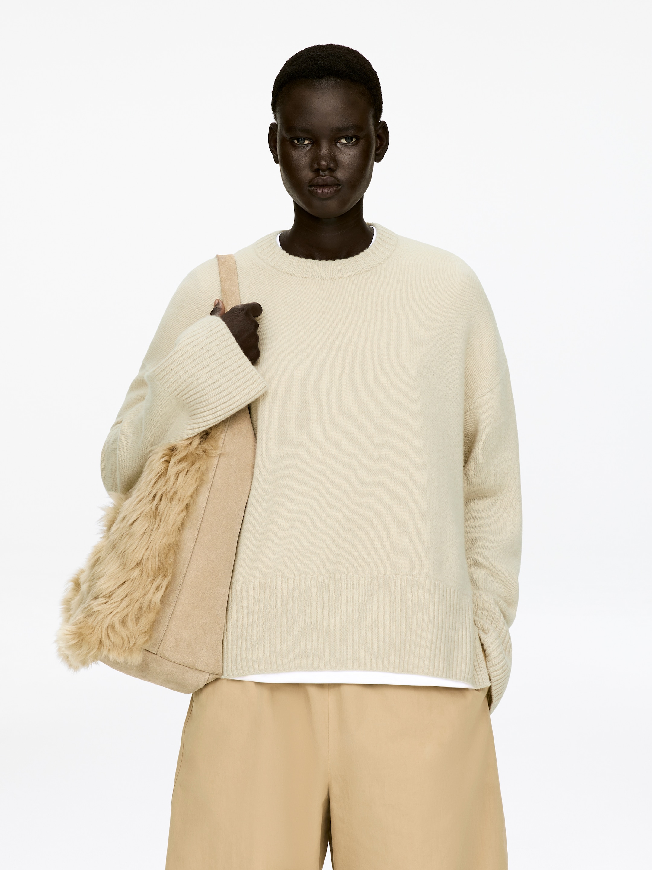 View larger image: A cream-colored, relaxed-fit jumper, featuring ribbed cuffs and a ribbed hem, is being worn by the model. A white t-shirt is visible underneath, and the model is carrying a beige bag with a faux fur panel against the jumper's side.