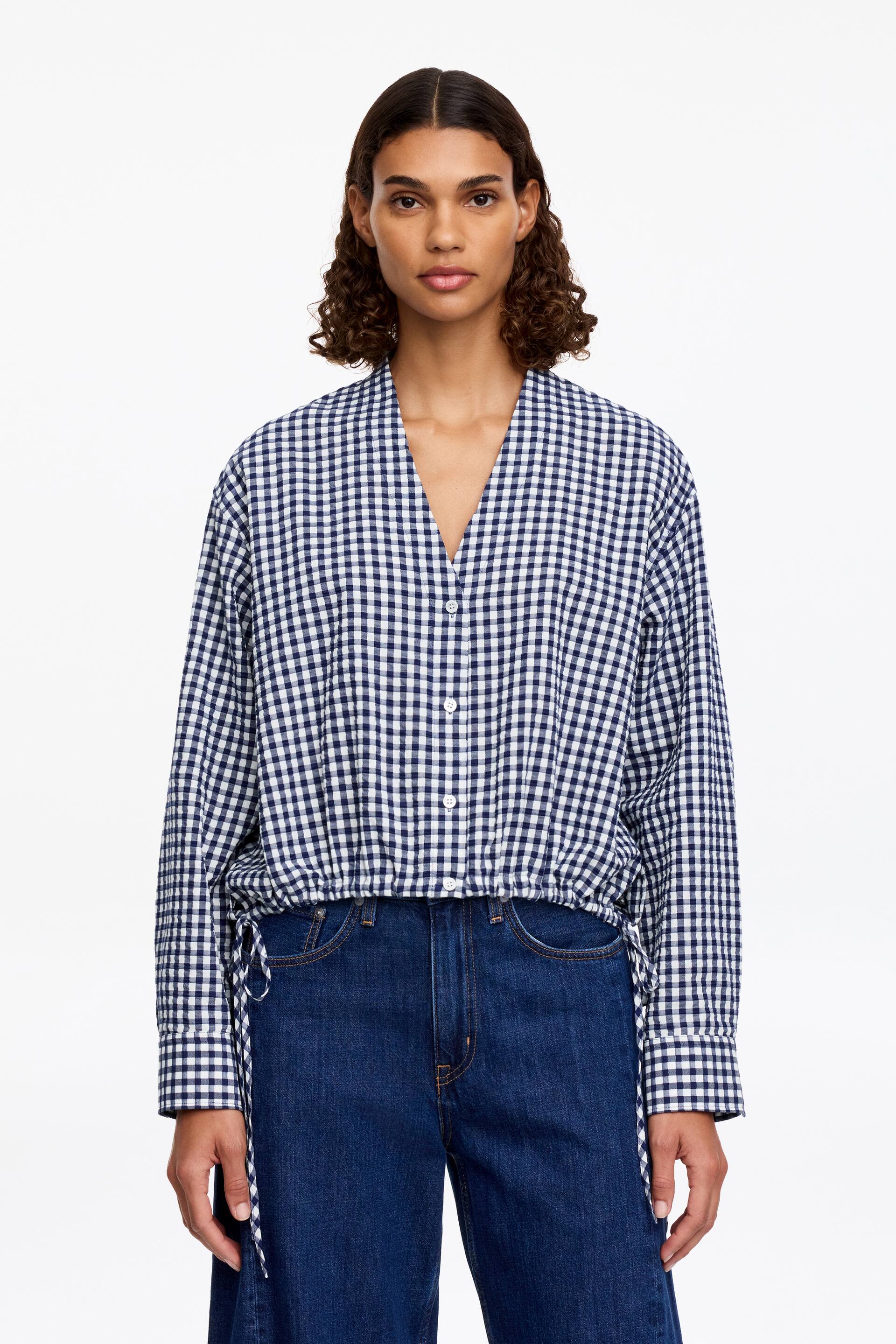 Ver imagem maior: Uma blusa de seersucker com decote em V e frente abotoada, finalizada com detalhes de cordão ajustável na bainha, está a ser usada pela modelo. Apresenta um padrão xadrez em azul e branco, com os cordões visíveis nas laterais, e está combinada com jeans azul escuro.