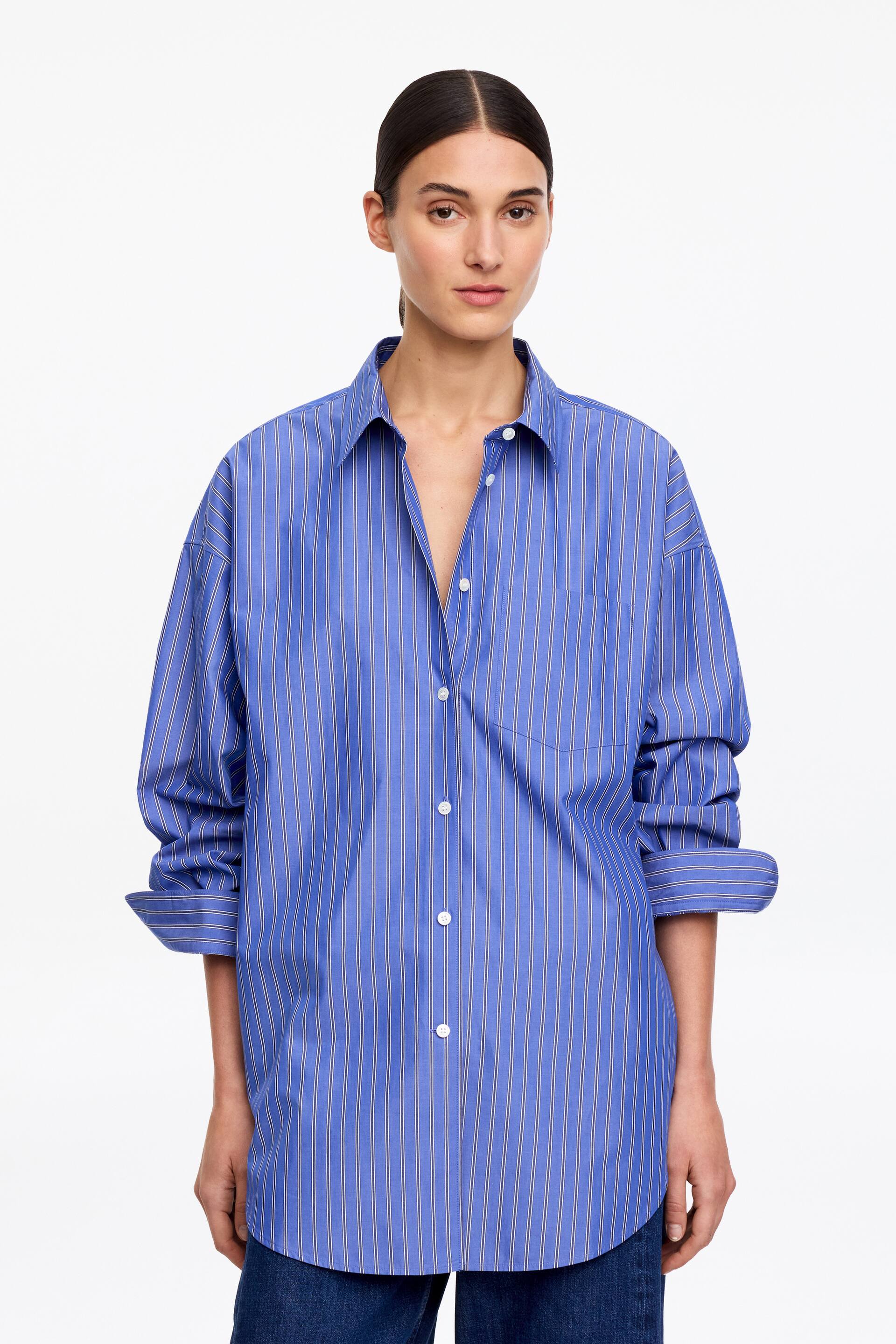 View larger image: The model is wearing an oversized blue shirt adorned with white vertical stripes, featuring a pointed collar, wide sleeves with rolled cuffs, and a single chest pocket. The shirt's top buttons are left undone, creating an open neckline, and the model is also wearing dark blue trousers.