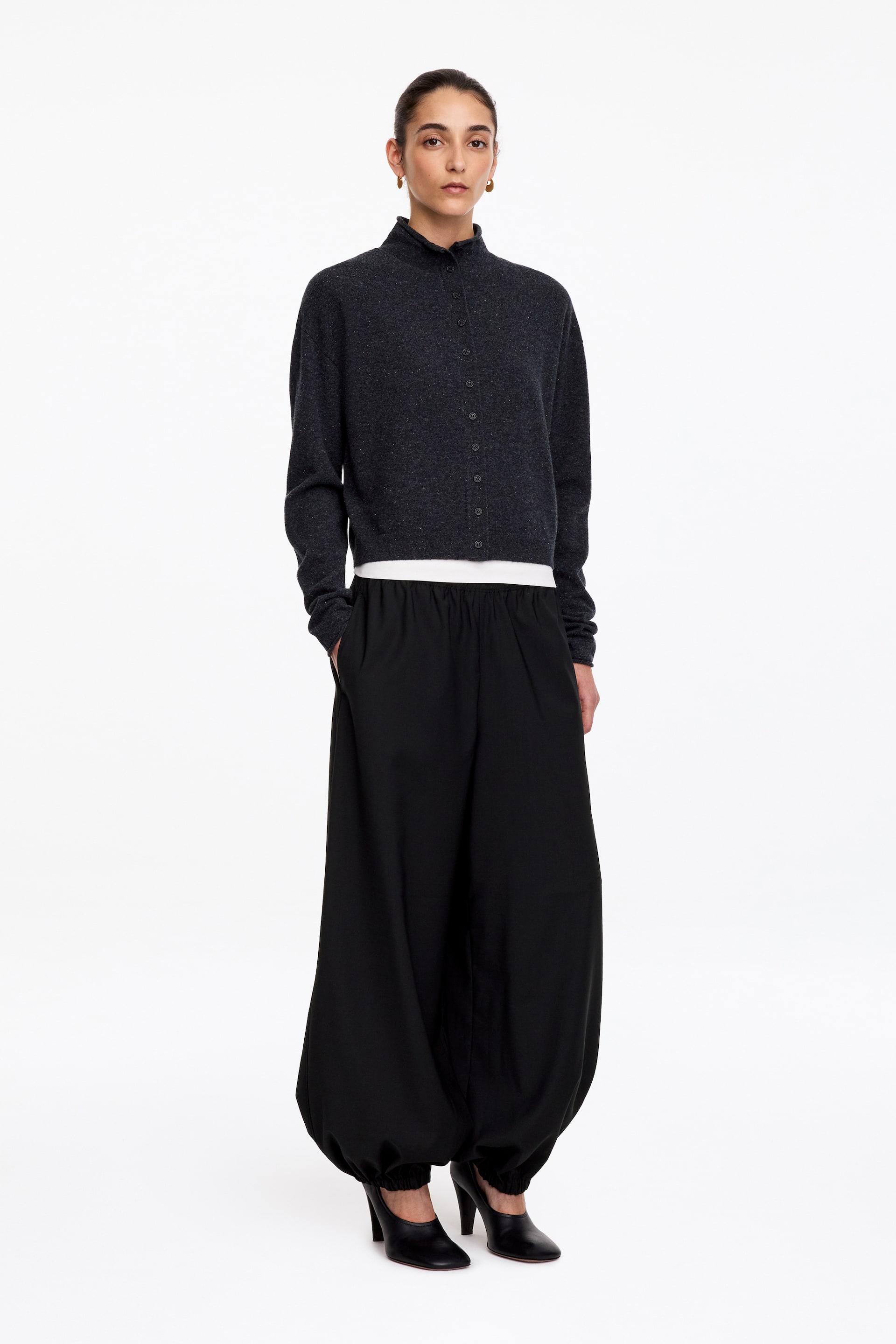 Größeres Bild anzeigen: Maßgeschneiderte schwarze Hose mit entspannter Barrel-Leg-Silhouette und elastischem Bund, getragen von dem Model, das mit einer Hand in einer Seitentasche steht. Die Hose läuft an den Knöcheln zu und verfügt über geraffte Beinabschlüsse; dazu werden eine dunkelgraue Strickjacke mit Knopfleiste und schwarze Schuhe mit Absatz kombiniert.