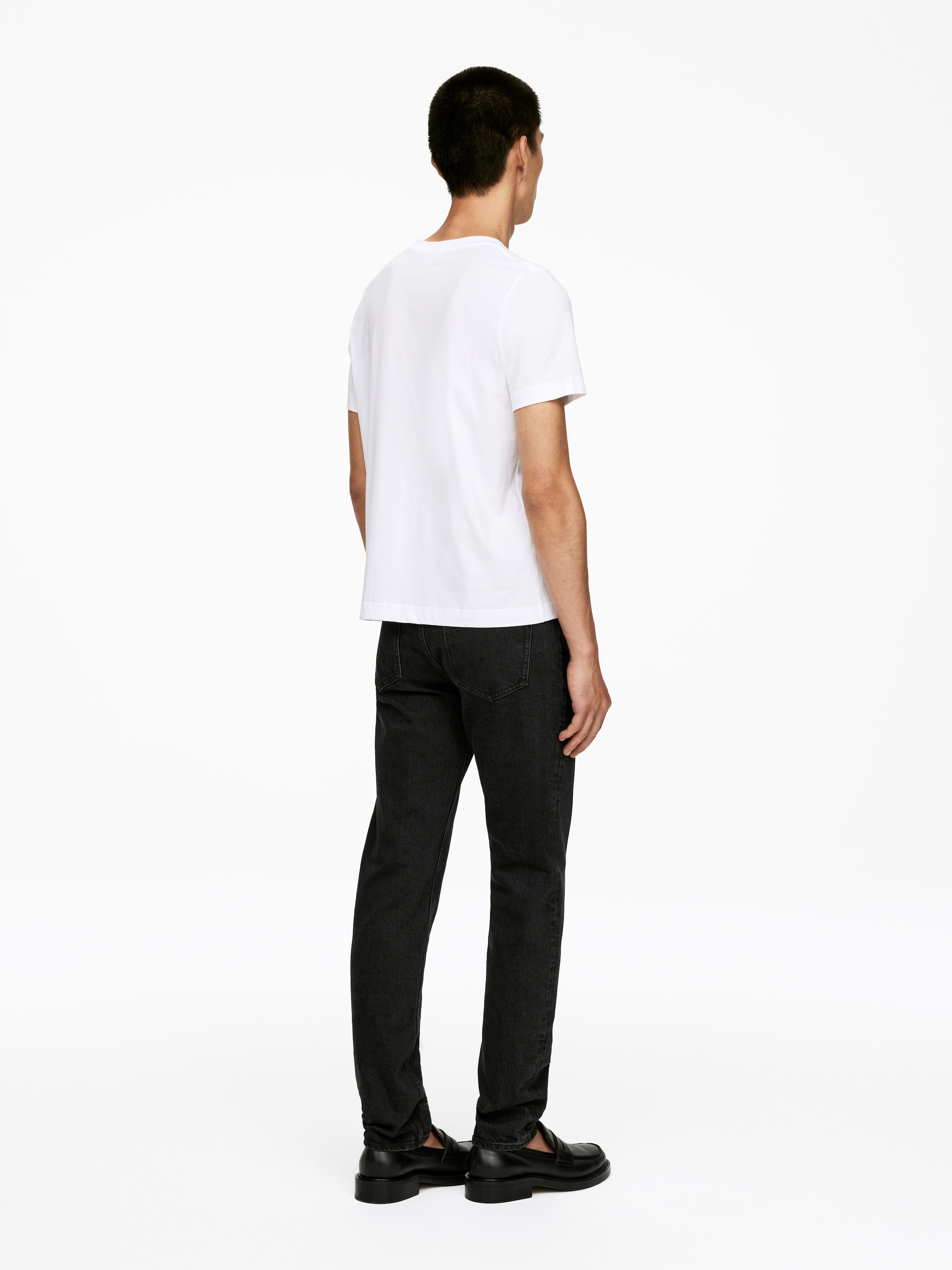 Ver imagem maior: Uma t-shirt branca lisa de gola redonda com mangas curtas e costas lisas está a ser usada, combinada com jeans pretos de lavagem escura com um corte direito e bolsos traseiros visíveis. Mocassins pretos com uma sola robusta completam o conjunto, tudo apresentado de um ângulo traseiro-direito para destacar o seu ajuste e design.