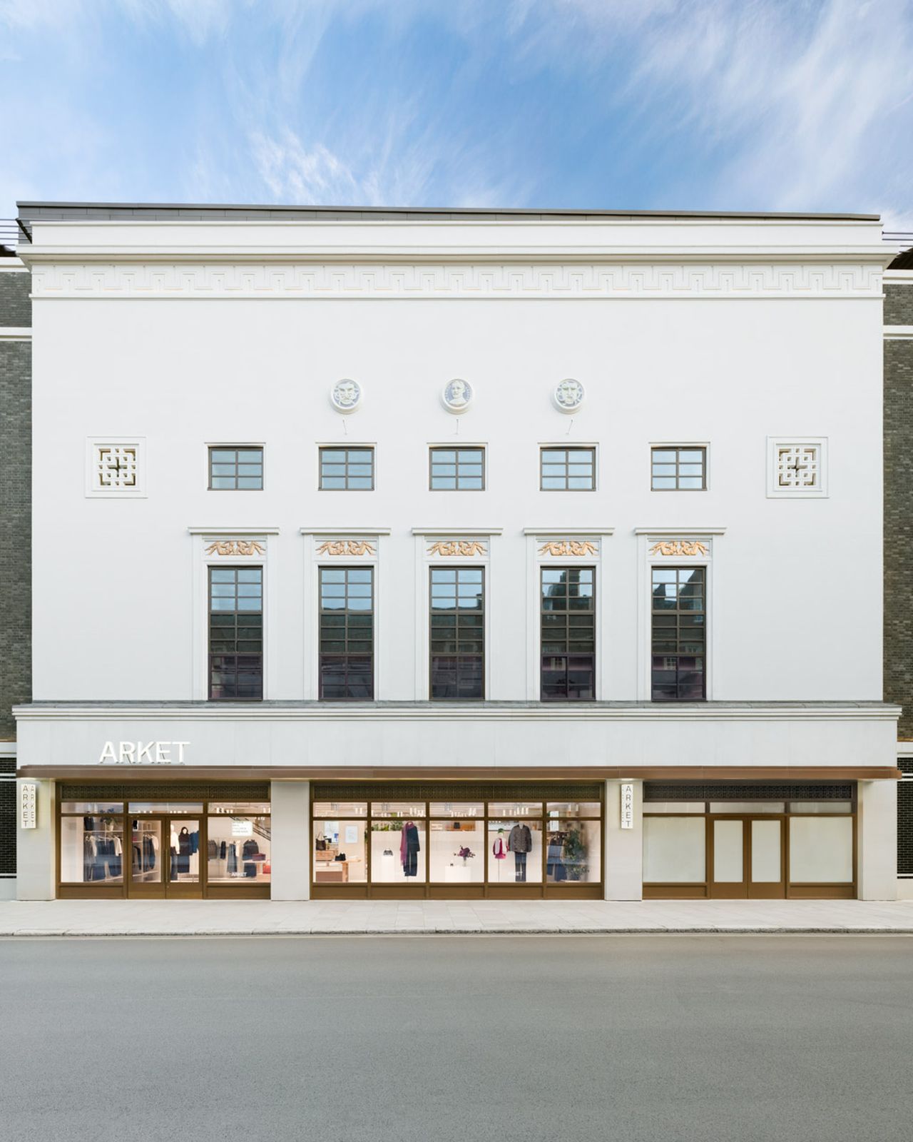 Front view of a modern Arket store with large windows displaying clothing, set in a white and gray building under a blue sky.