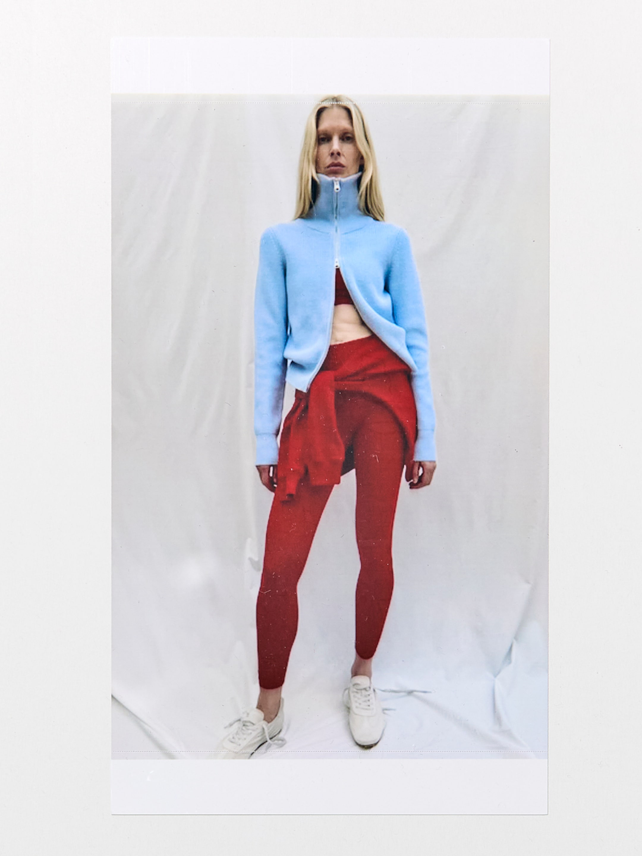 A person in a blue zip-up sweater over a red sports top, red leggings, sneakers, and a tied-around red sweater stands against a white backdrop.