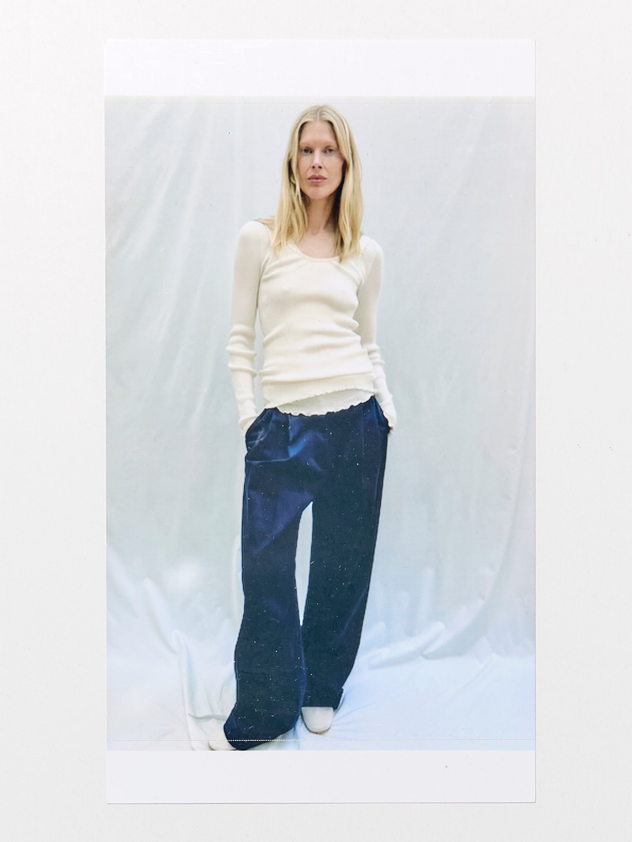 A person with long blonde hair wearing a white long-sleeve top and loose blue trousers, standing against a light-gray backdrop.