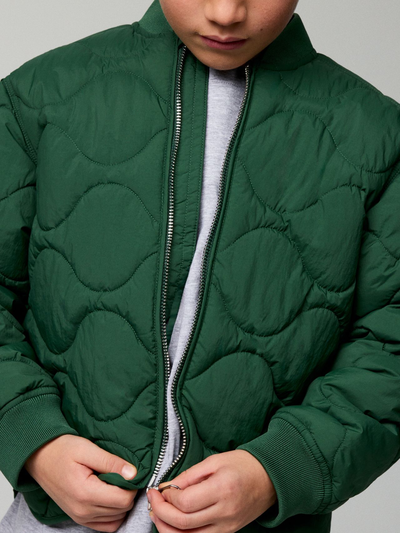 Person in a quilted green jacket, partially zipping it up over a gray shirt, with hands near the zipper.
