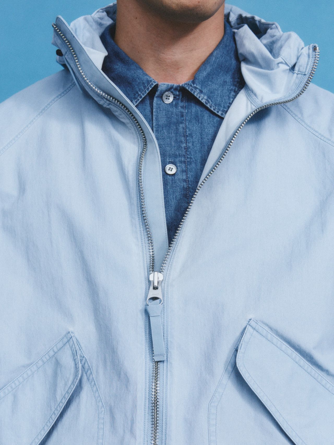 Close-up of a person wearing a light blue jacket over a denim shirt against a blue background. The jacket is unzipped slightly.