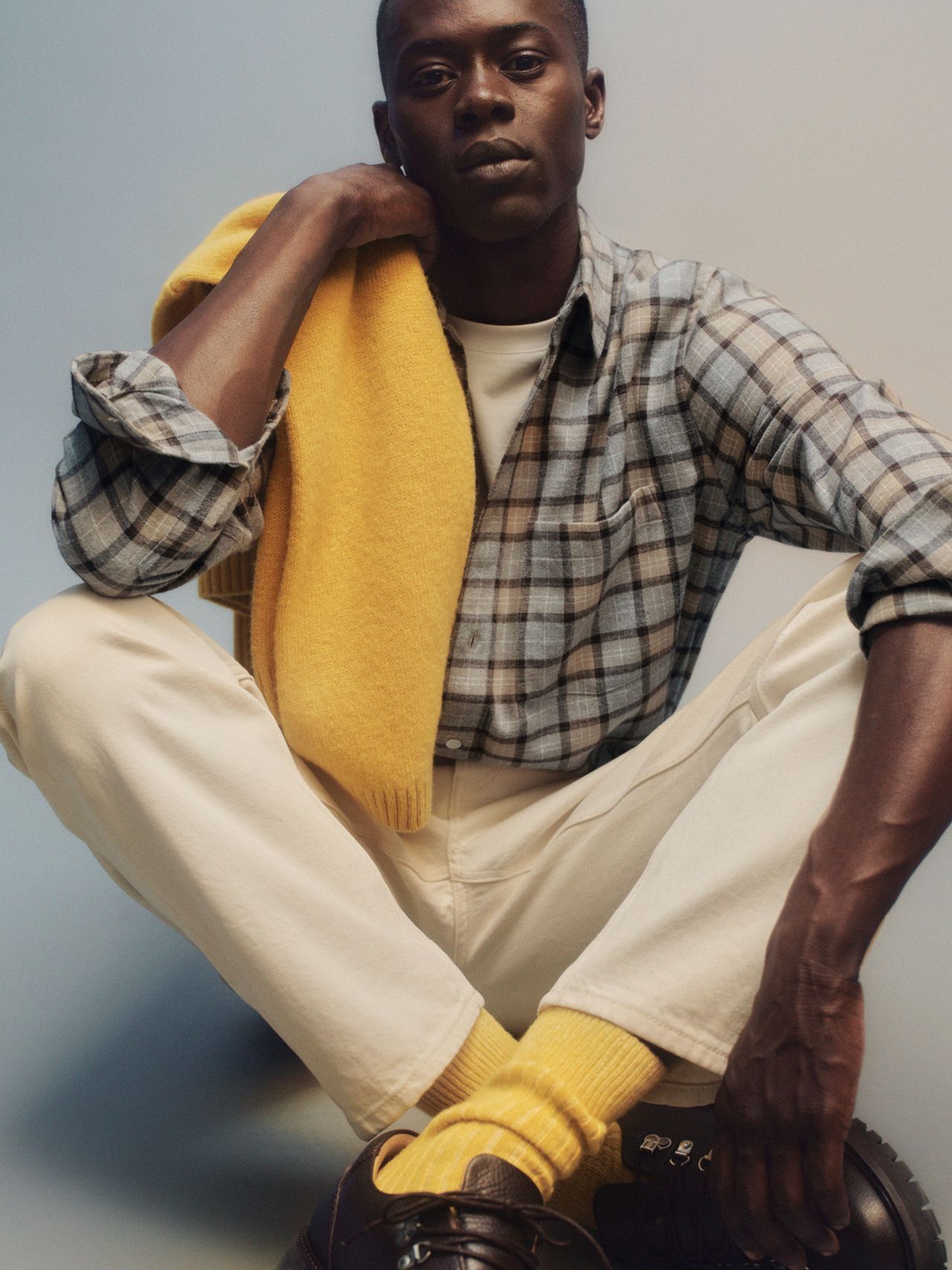 Seated man in plaid shirt and cream pants, yellow scarf draped over his shoulder, matching yellow socks and brown shoes.