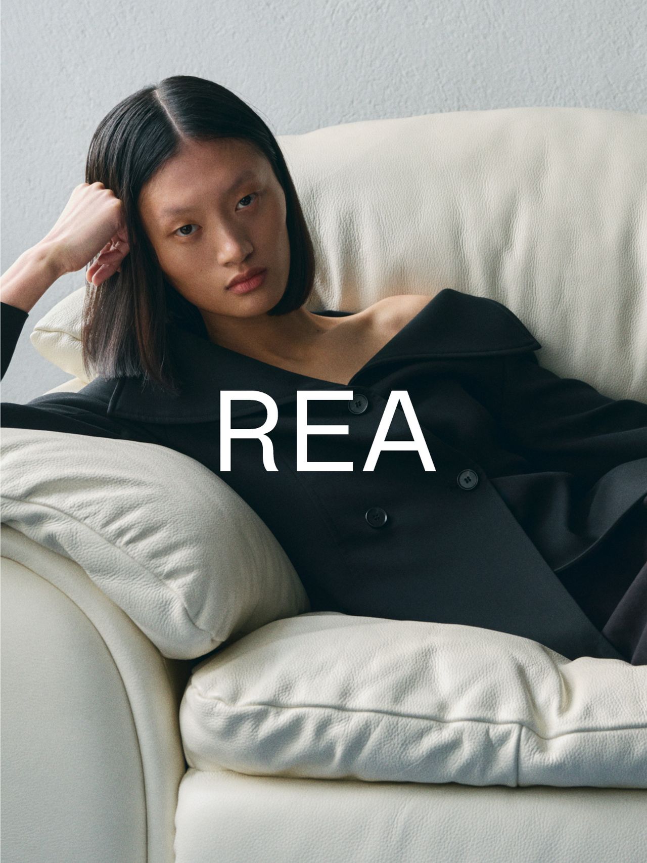 Person with straight hair in a black coat reclining on a white sofa, with the word "REA" overlaid on the image.
