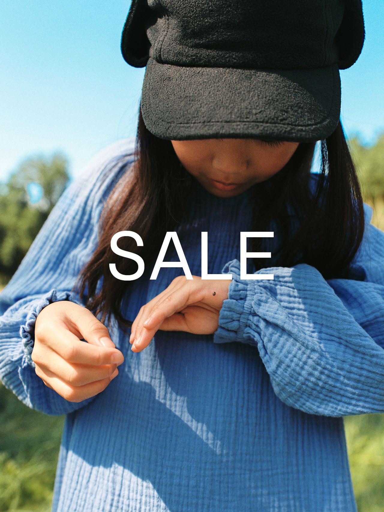 Child in a blue dress and black cap looks at their hand outdoors, with "SALE" text overlayed.