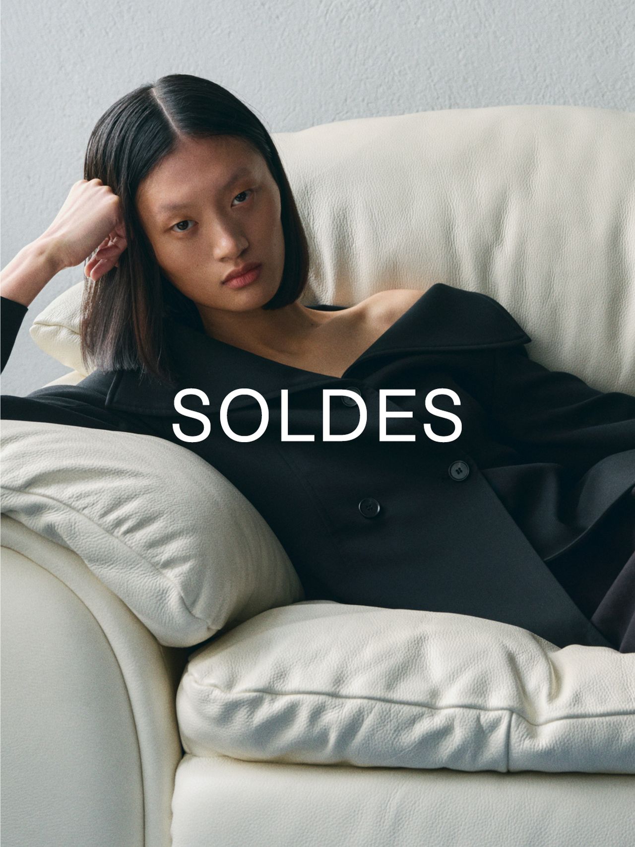 Person with straight hair reclines on a white sofa wearing a dark coat. The word "SOLDES" is overlaid on the image.