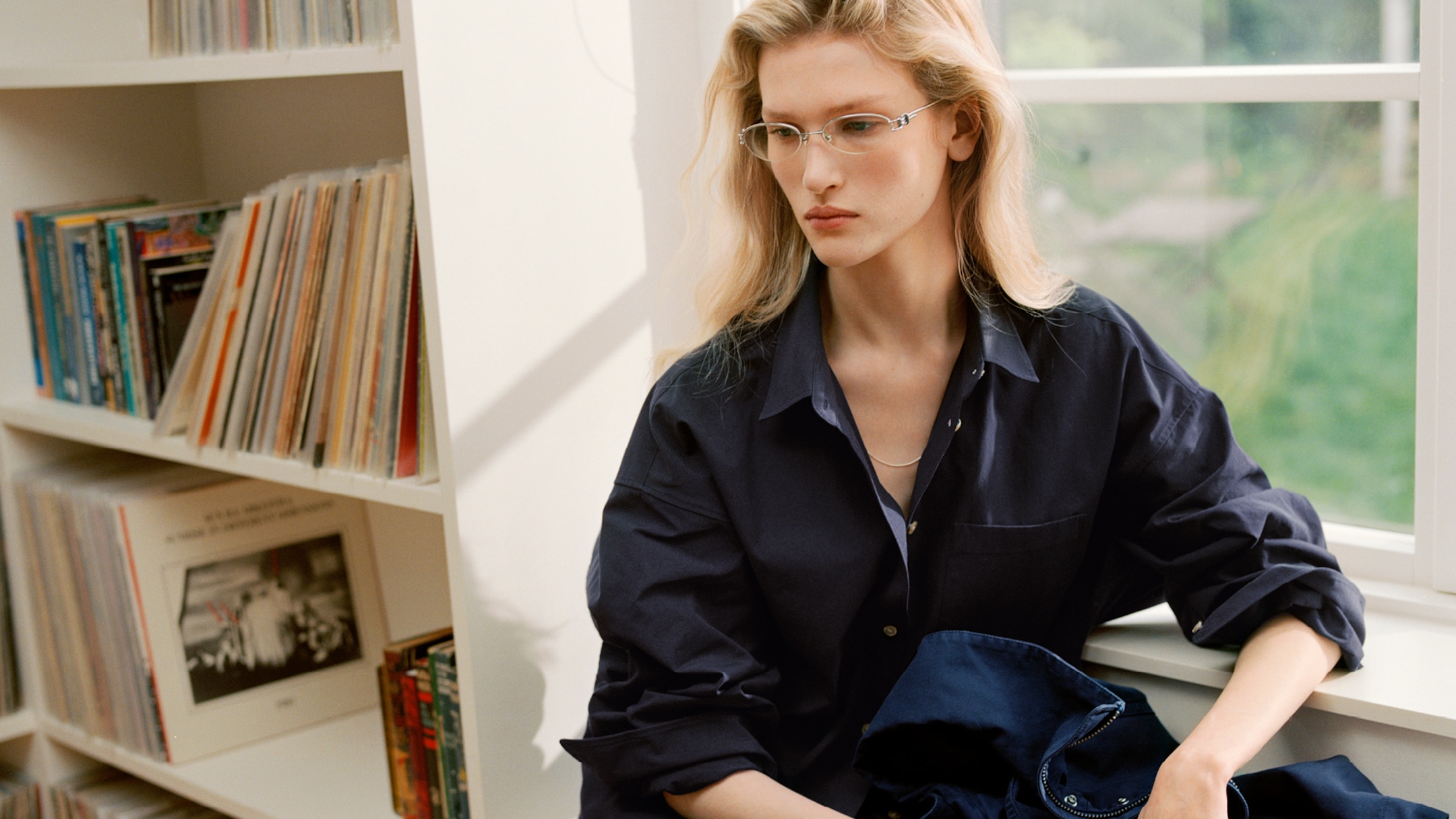 Woman with glasses sitting by a window, holding a folded navy shirt, with a shelf of records and books visible in the background.