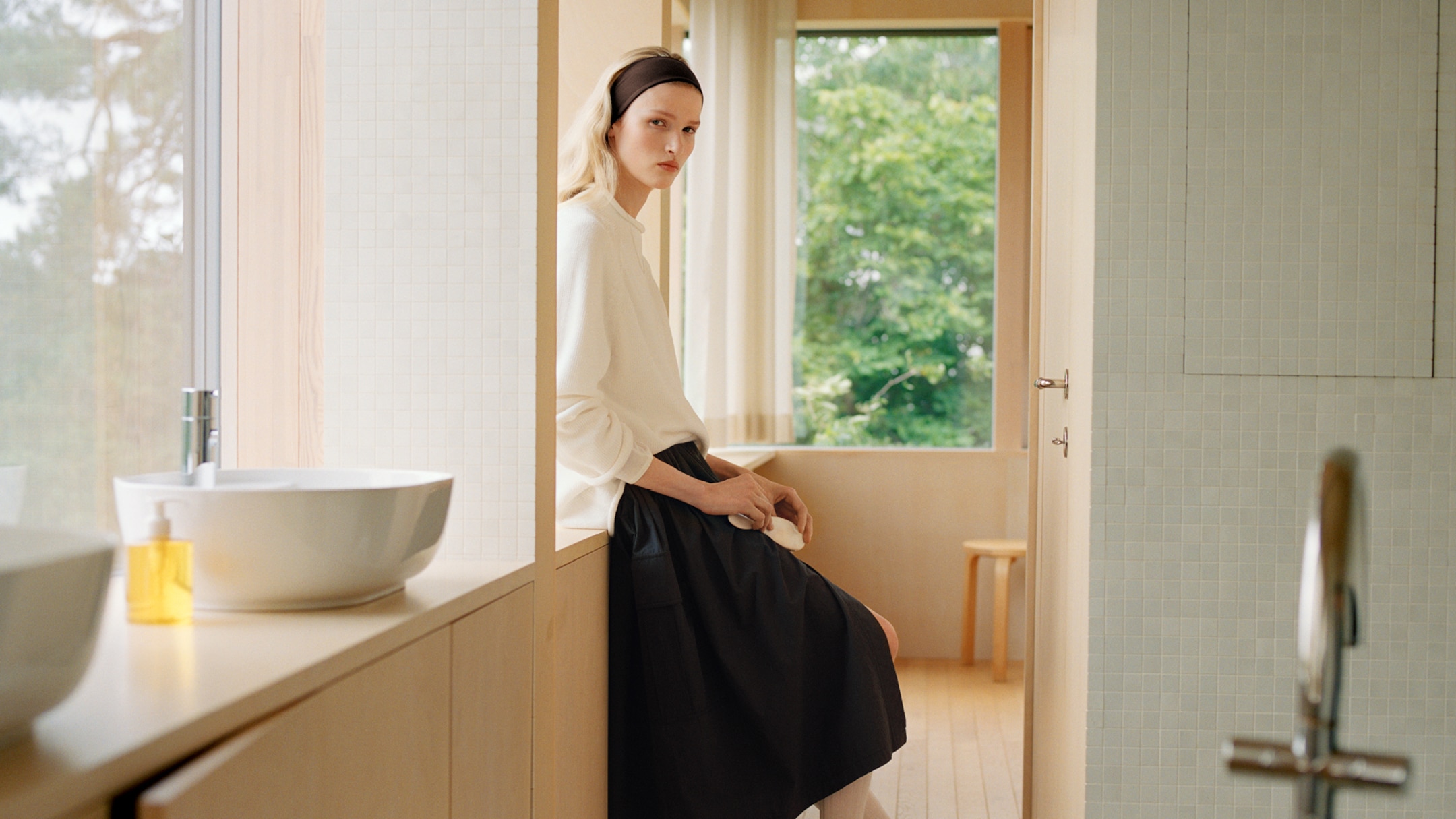 A person with a headband sits on a bathroom counter, dressed in a white top and black skirt, with a large window and greenery in the background.