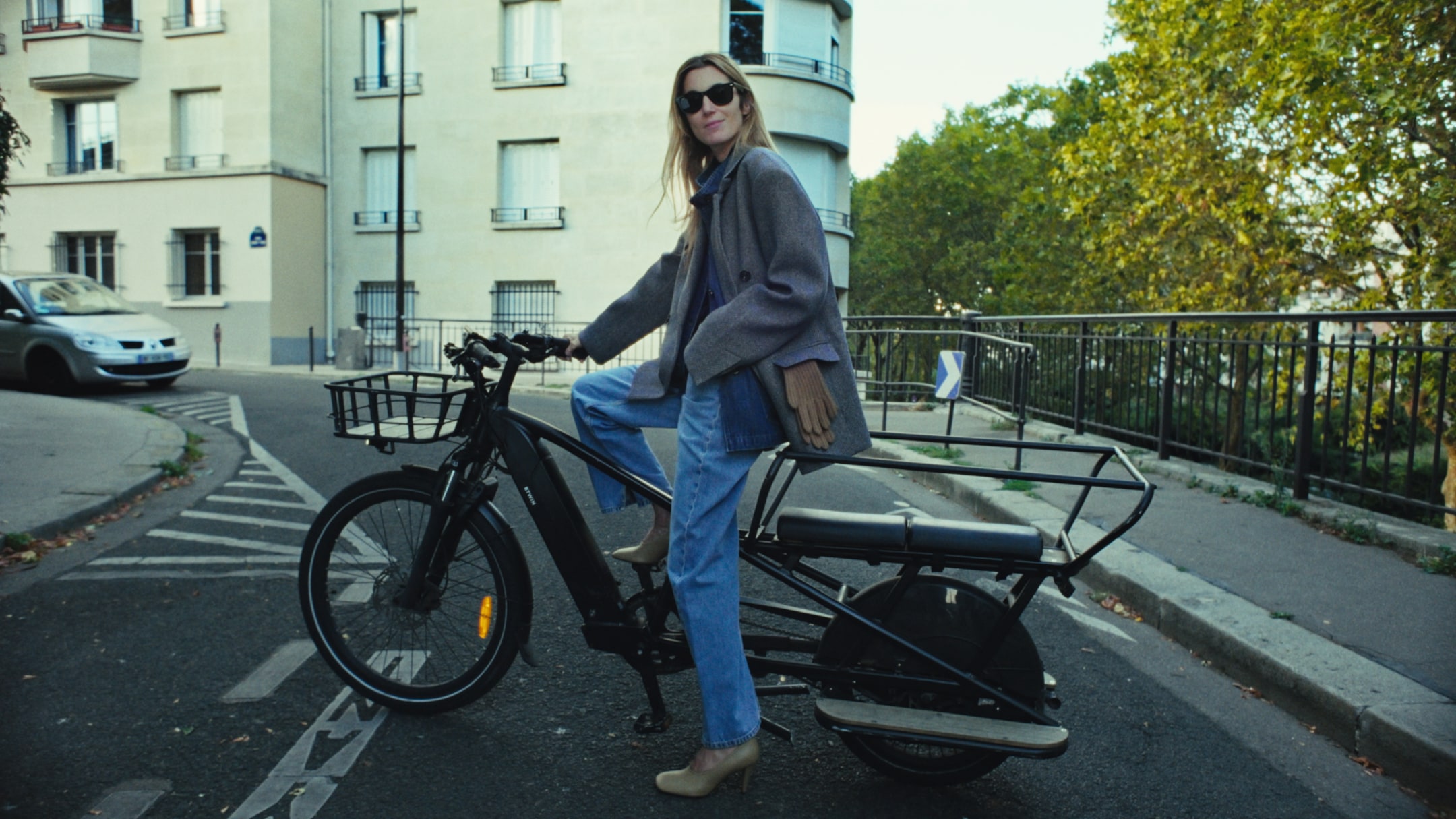 A person wearing sunglasses and a coat sits on a parked cargo bicycle on a city street, with buildings and trees in the background.